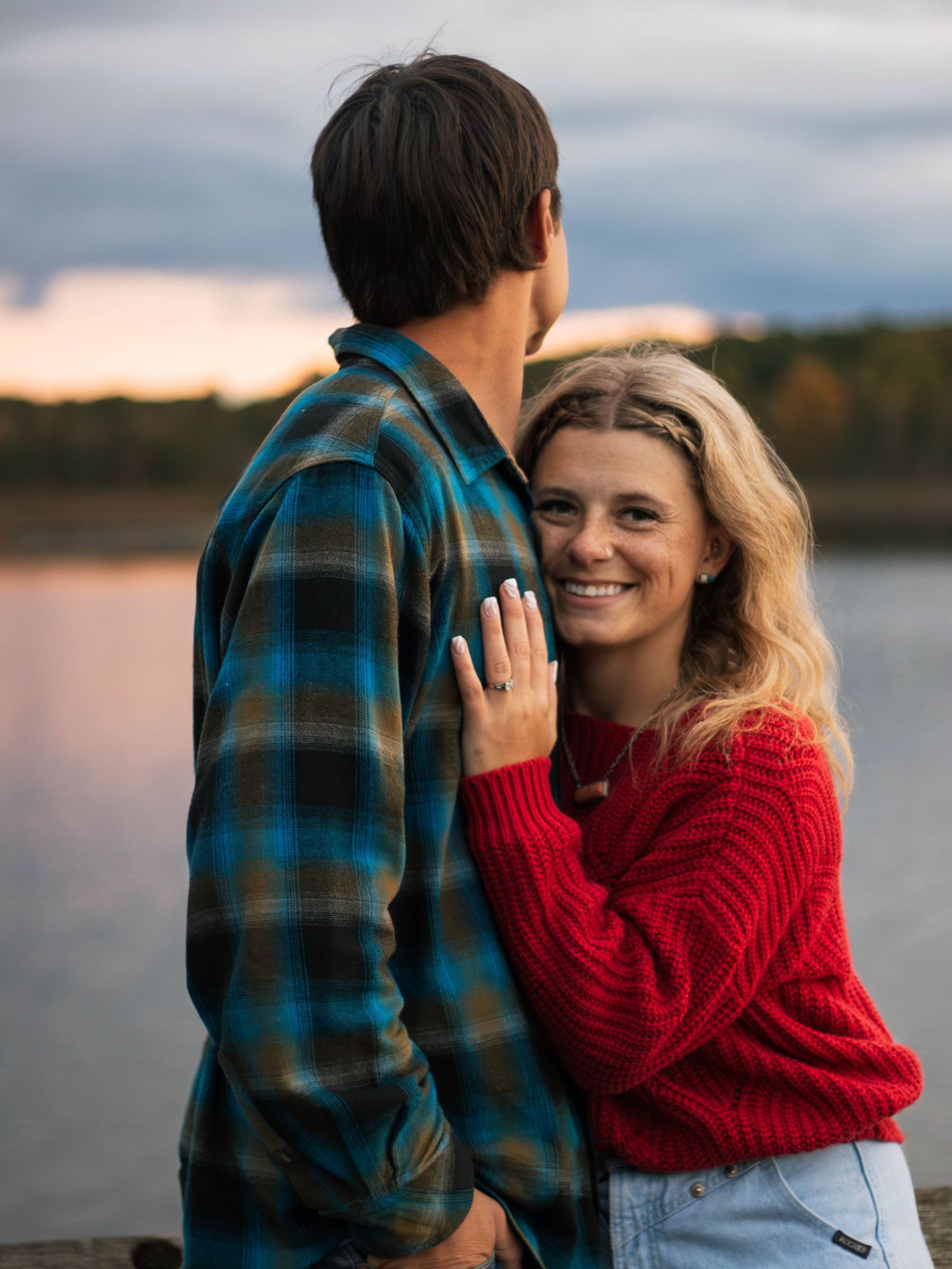 Couple sharing an intimate moment by a lake during sunset with person wearing red sweater.