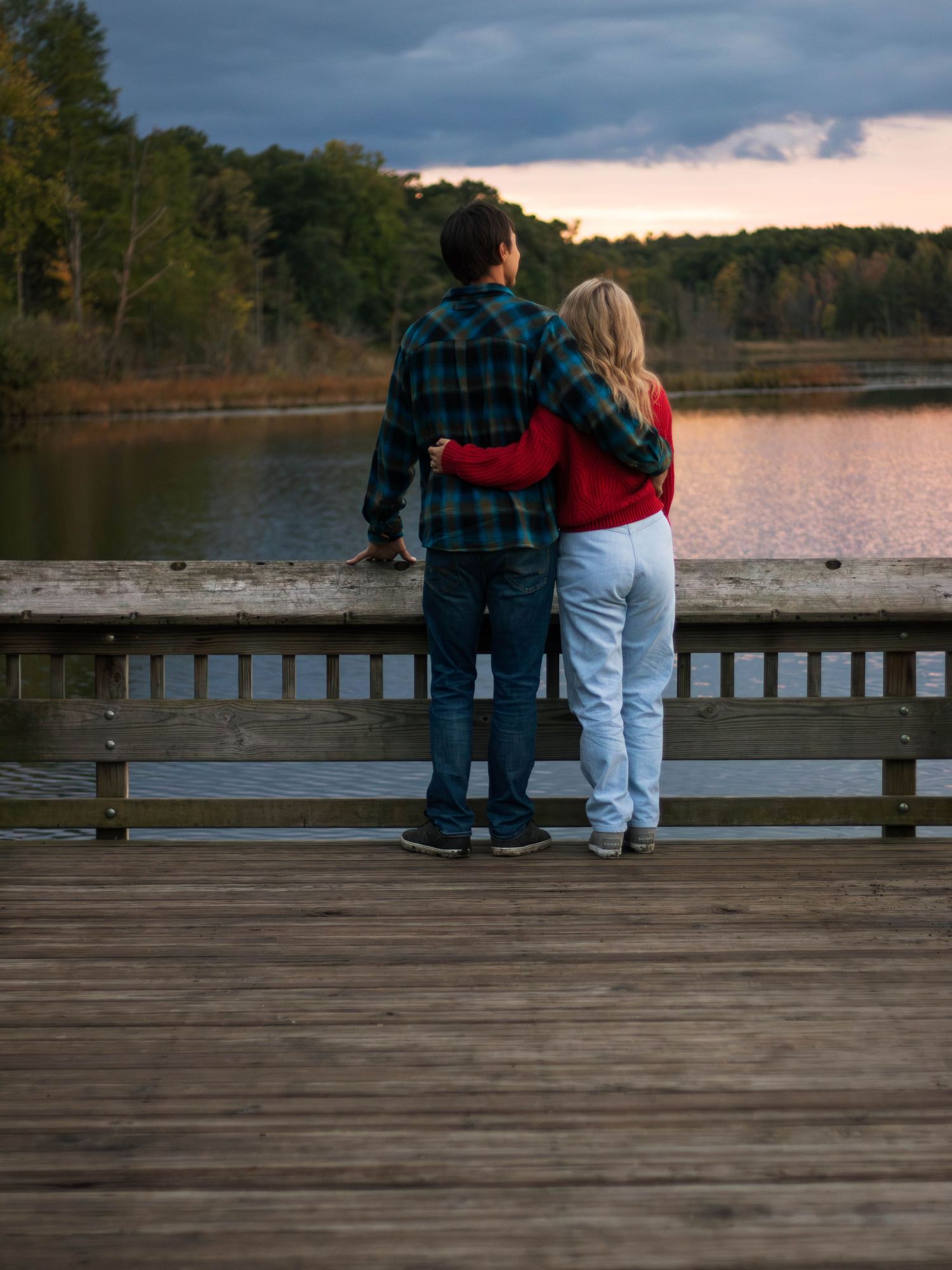 A couple stands close together on a wooden dock overlooking a lake at sunset.