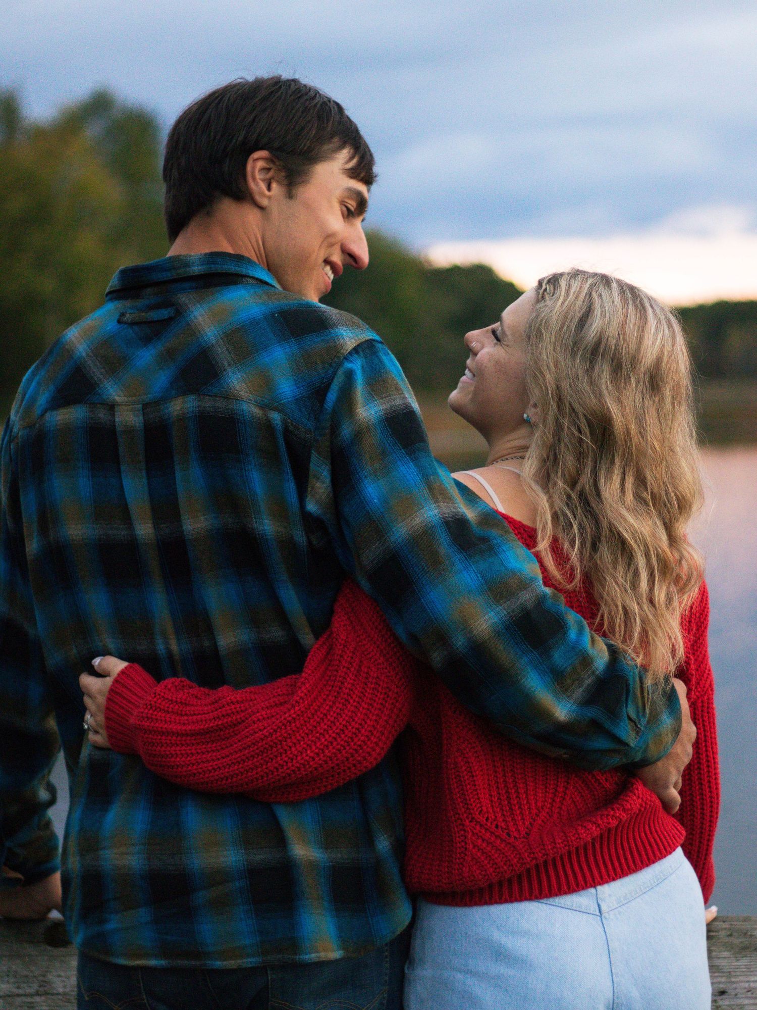 A couple in casual wear shares an intimate moment on a lakeside dock during sunset.