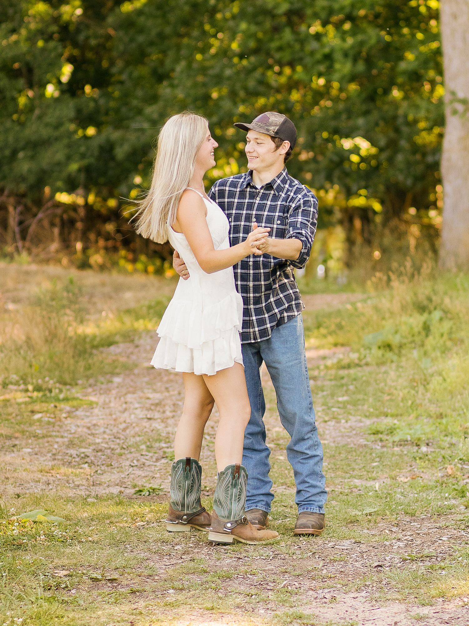 Couple in casual attire standing on dirt path with autumn trees in background.