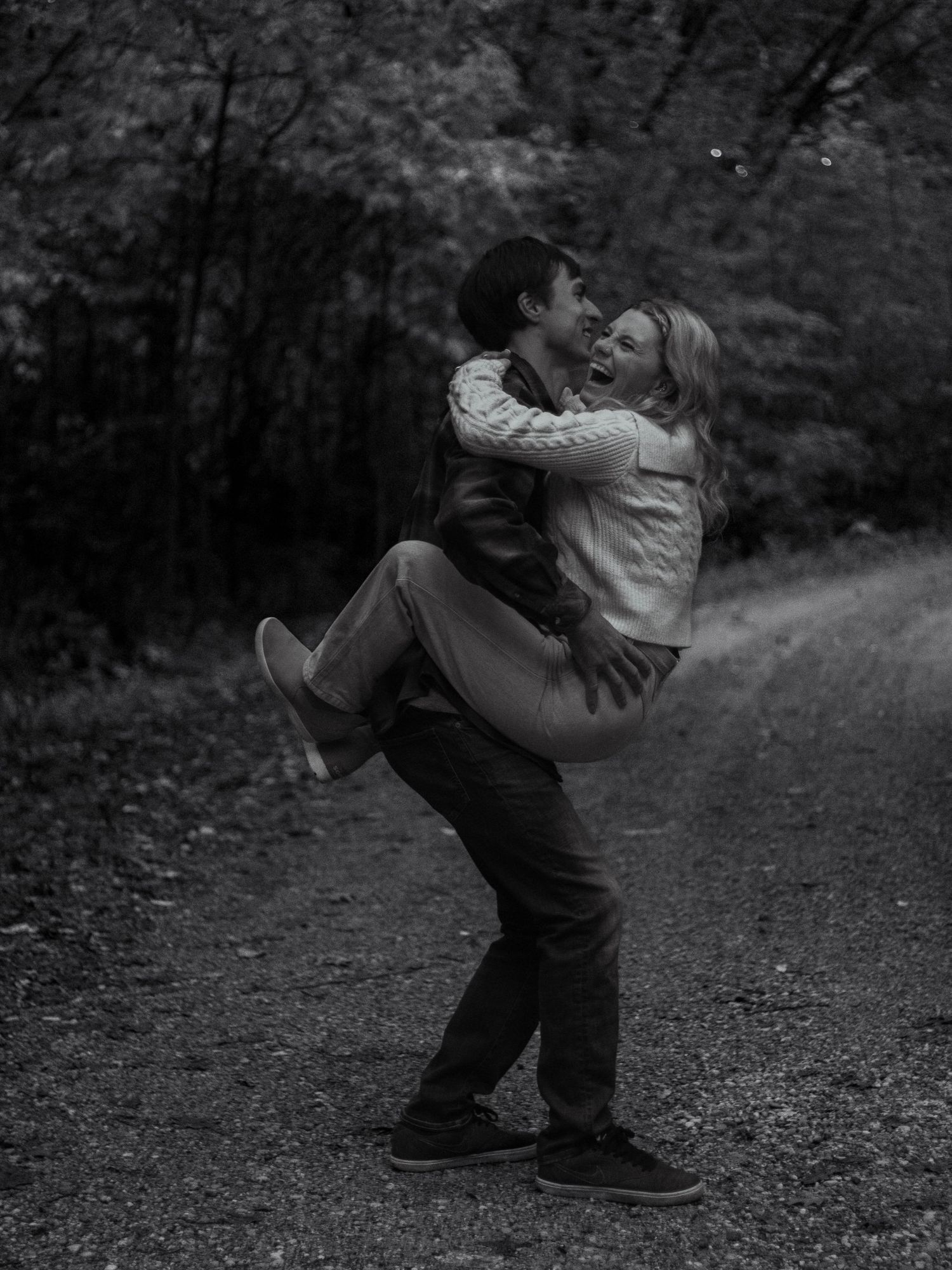 Black and white photo of couple sharing playful moment on forest path.