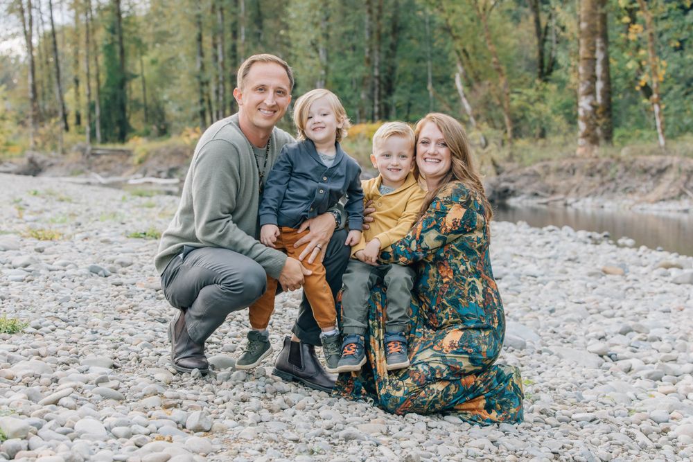 Family of four smiling together on a rocky riverbank surrounded by autumn trees.