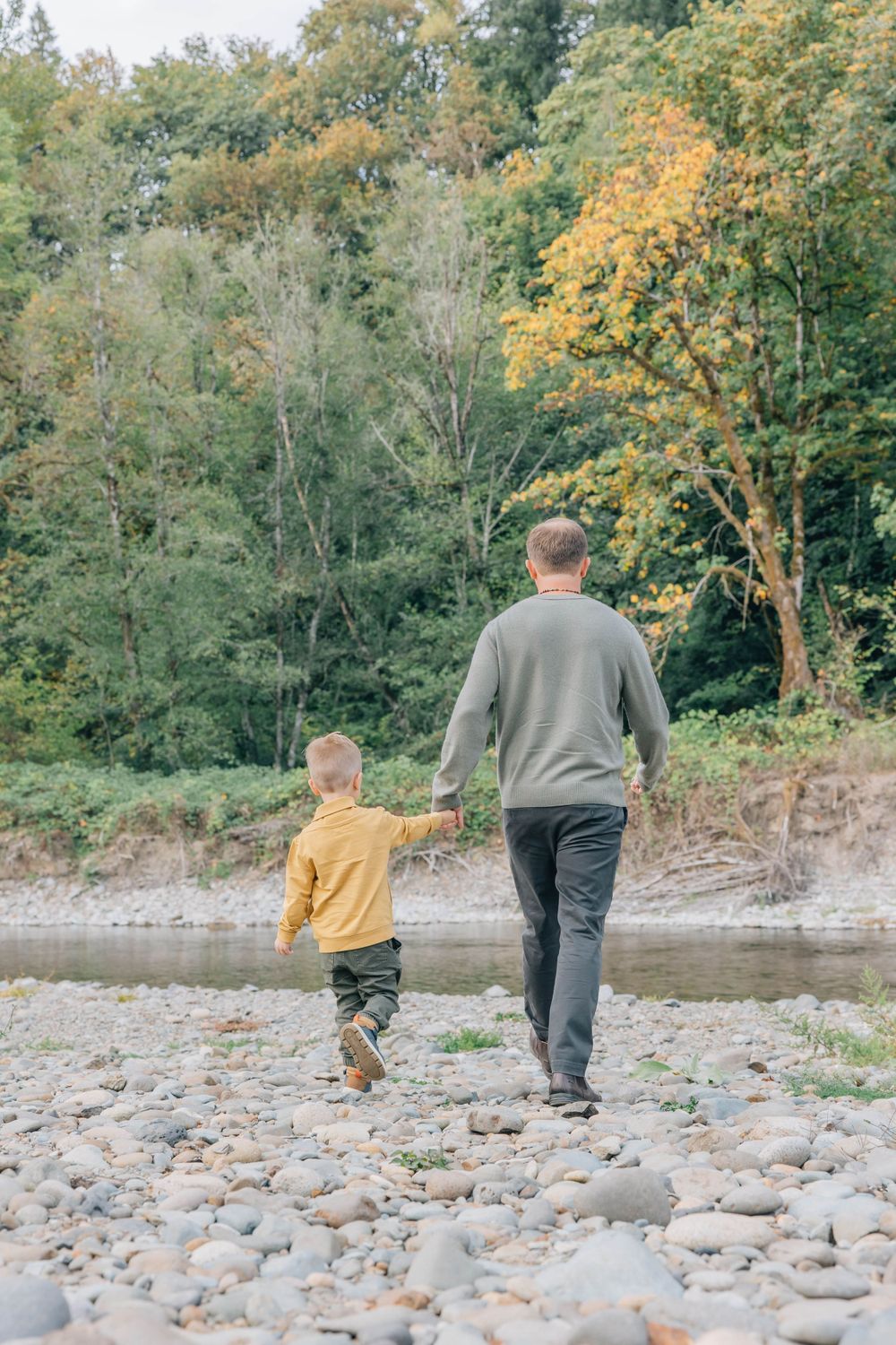 Father and young son walking hand in hand along a rocky riverbank in autumn.