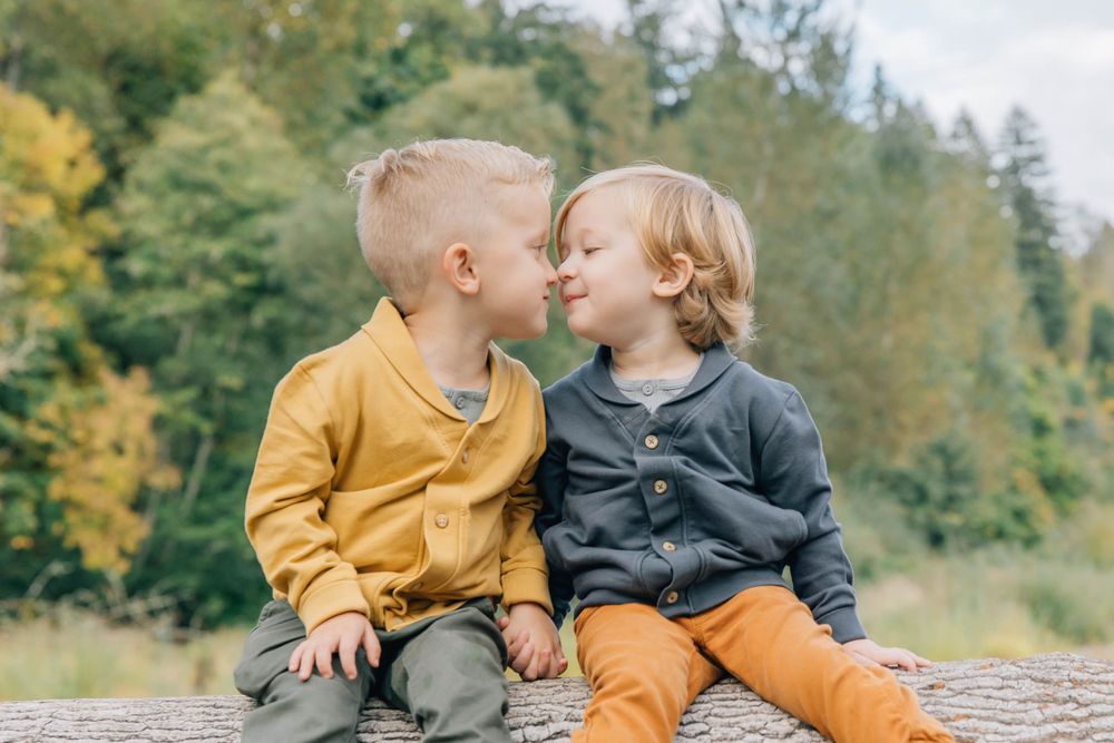 Two young brothers sitting on a log touching noses affectionately in autumn forest.