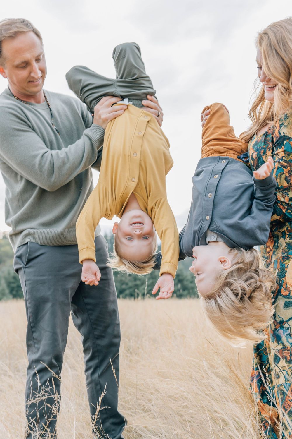 Parents playfully hold two young boys upside down in a golden grass field outdoors.