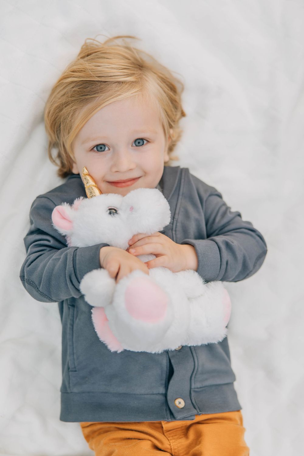 Adorable blonde toddler smiles while hugging a white unicorn stuffed animal toy.