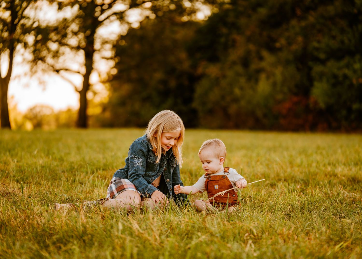 Bald grandfather crouches beside smiling baby in brown overalls in golden sunset field.