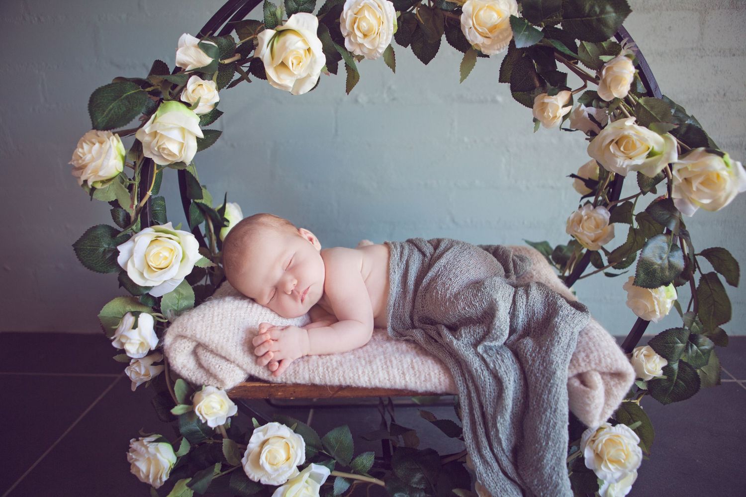 A sleeping newborn nestled in soft pink and grey fabric surrounded by delicate cream-colored roses.