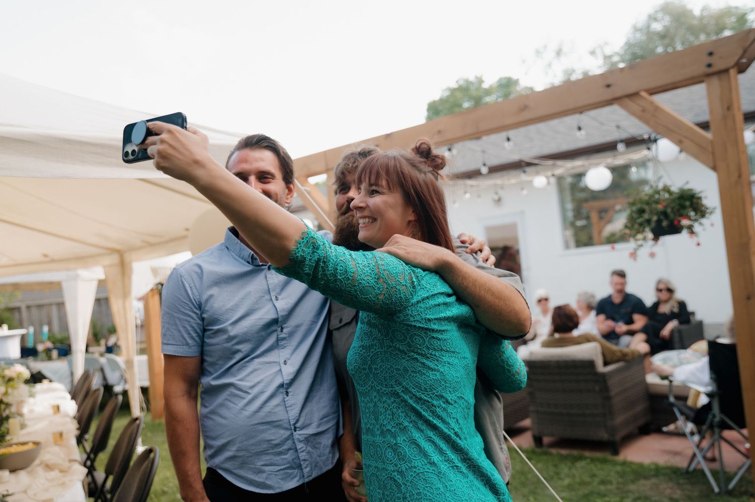 A woman in a teal dress takes a selfie with a man at an outdoor backyard party with string lights.