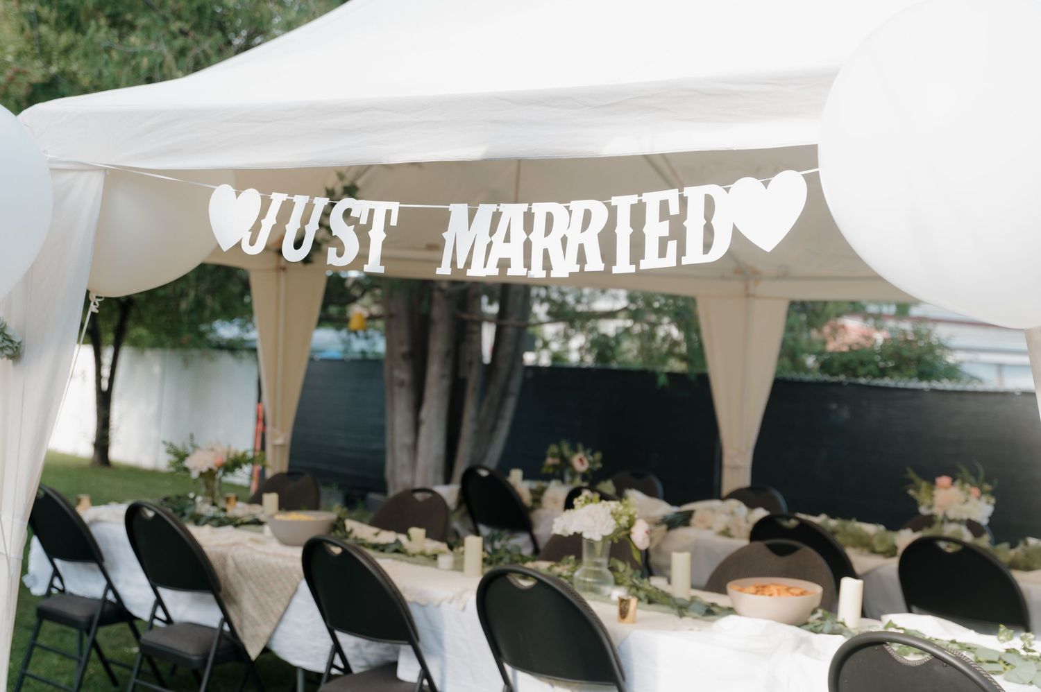 Just Married banner hanging under a white tent at an outdoor wedding reception with decorated tables and black chairs.