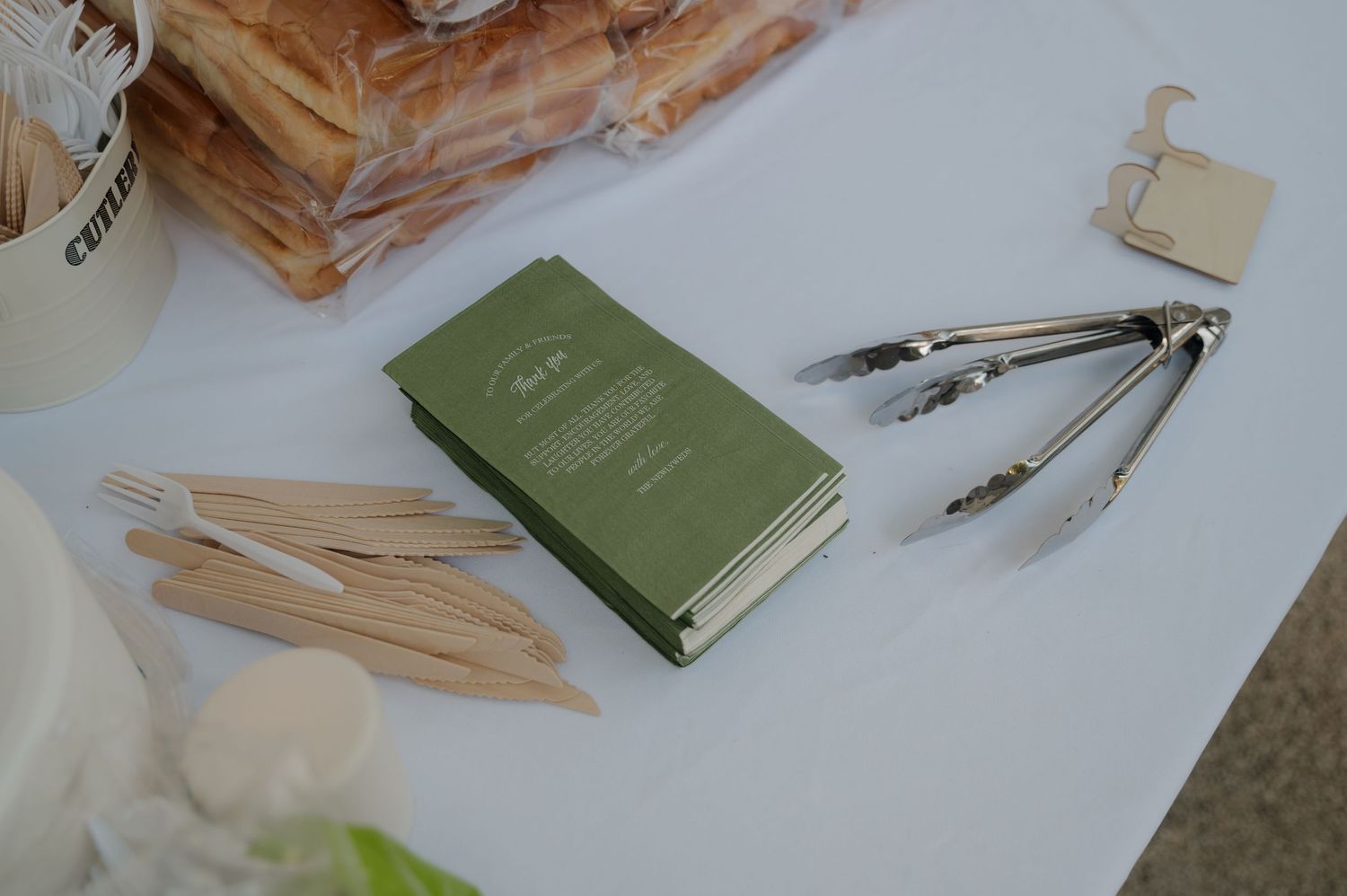 Green notebook with wooden cutlery and metal tongs on a white table at an outdoor event.