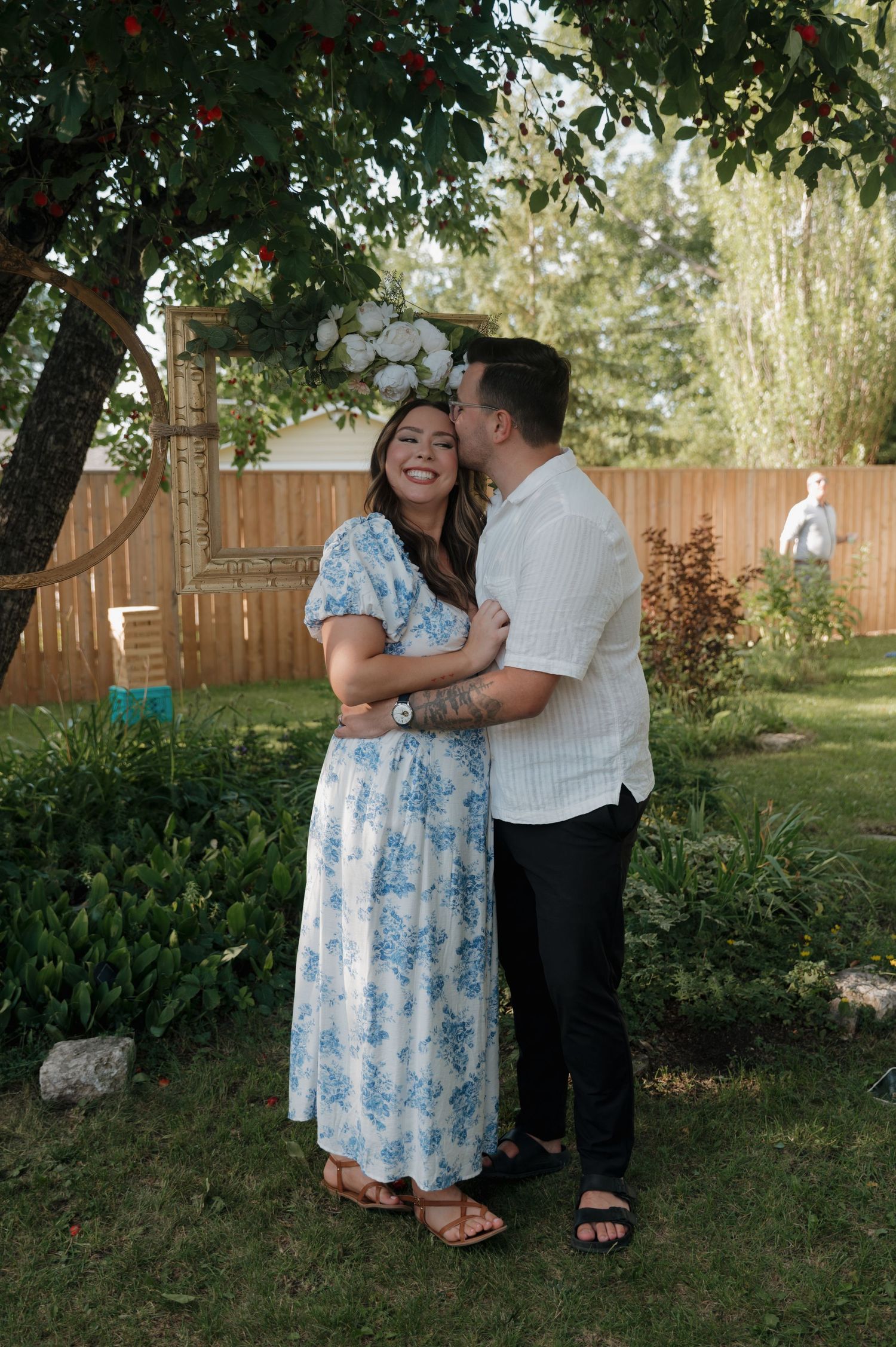 Couple embracing and smiling in a lush backyard garden, woman wearing a blue floral dress with a flower crown.
