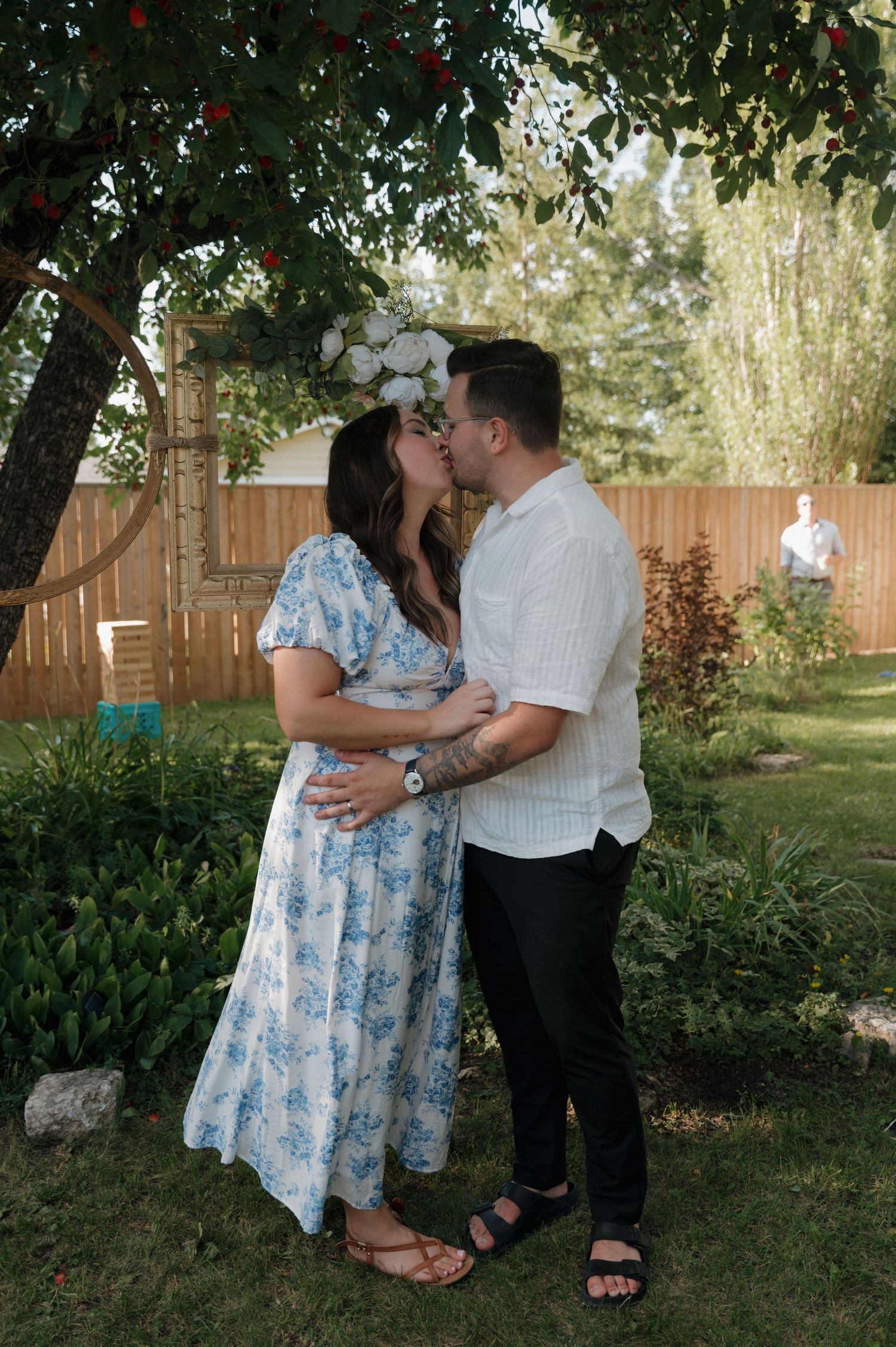 Couple sharing a romantic kiss in a lush backyard garden, woman in floral blue dress with flower crown.