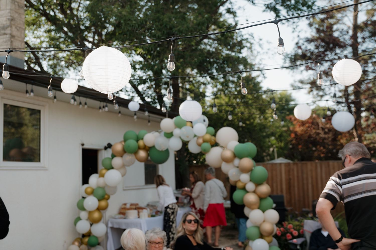 Colorful green, white, and gold balloon arch decorates an outdoor backyard party with string lights.