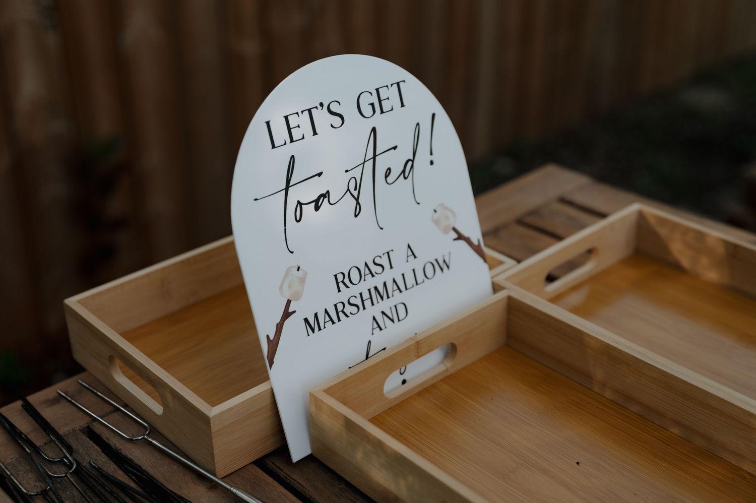 A decorative wedding sign reading Let's Get Toasted Roast a Marshmallow on a wooden tray.