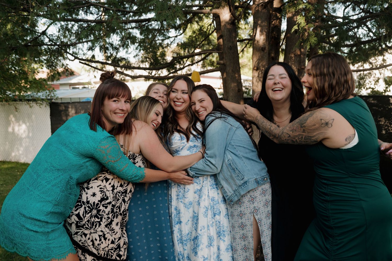 Group of women hugging and laughing together outdoors at a wedding celebration under trees.