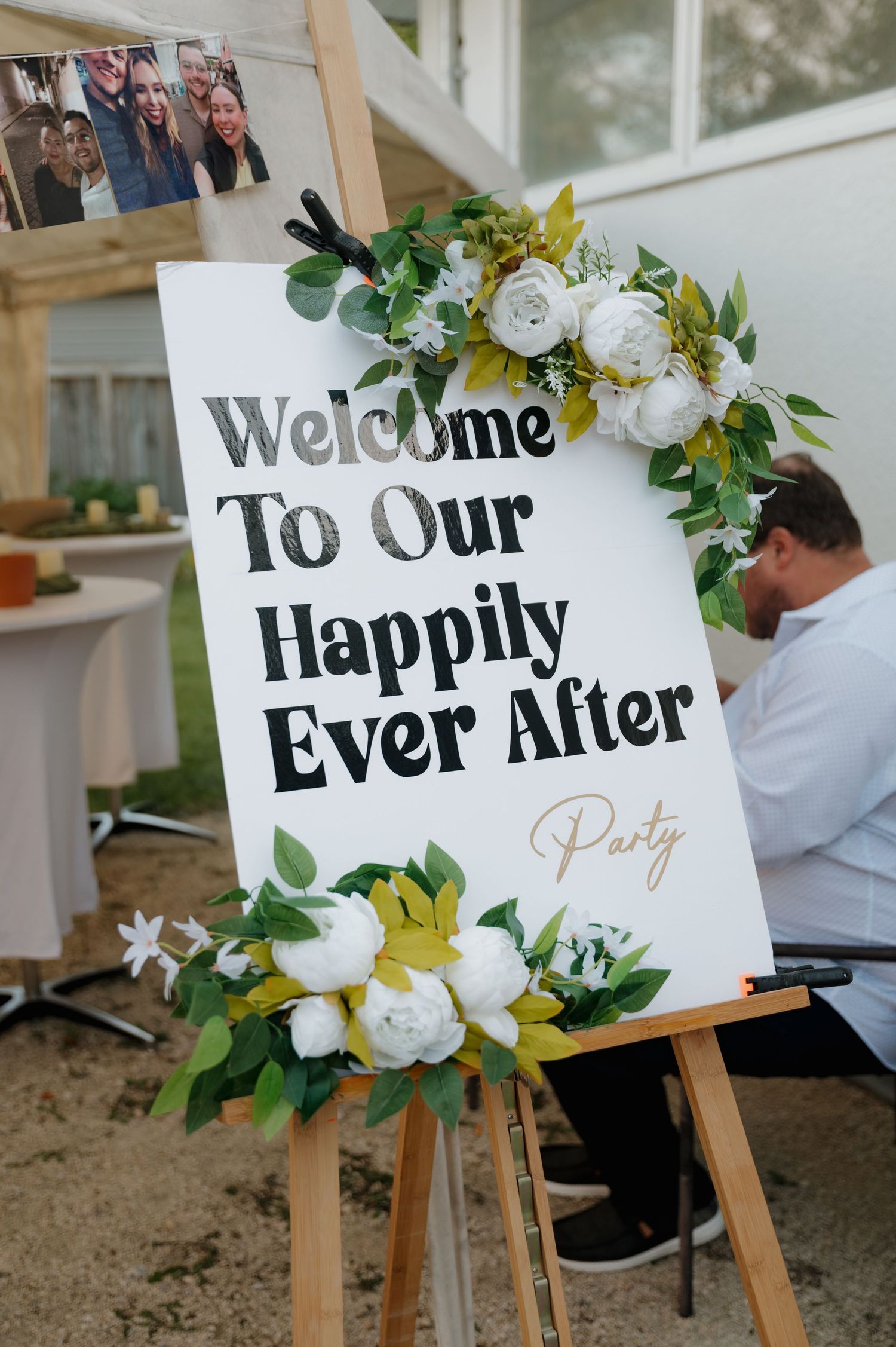 Wedding welcome sign reading Happily Ever After decorated with white roses and greenery on a wooden easel.