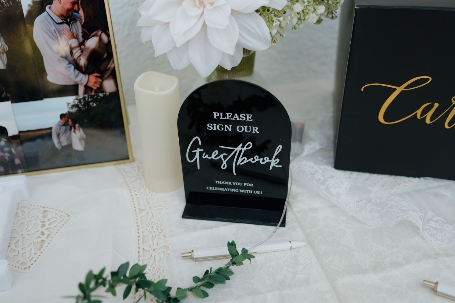 Black acrylic wedding guestbook sign on a decorated table with candle, greenery, and floral arrangements.