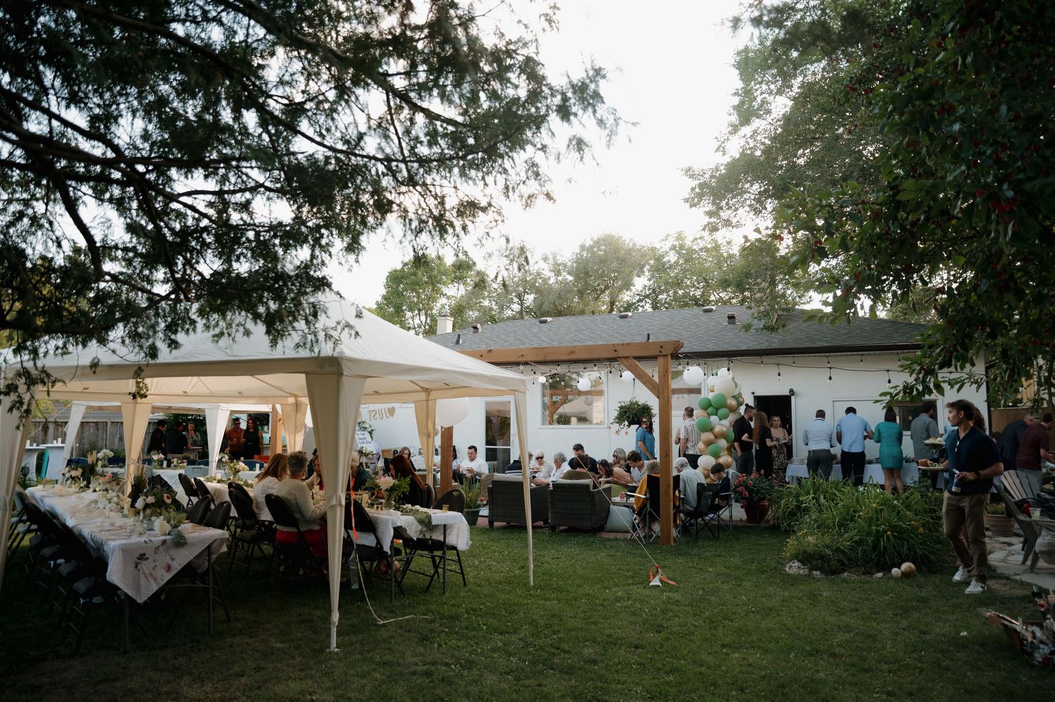 Outdoor backyard wedding reception with white tent, string lights, and guests mingling.
