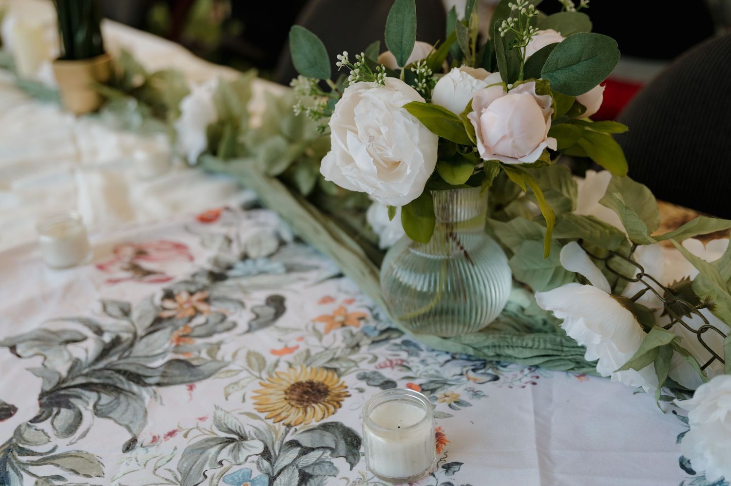 Elegant floral centerpiece with white roses and greenery in a glass vase on a patterned tablecloth.