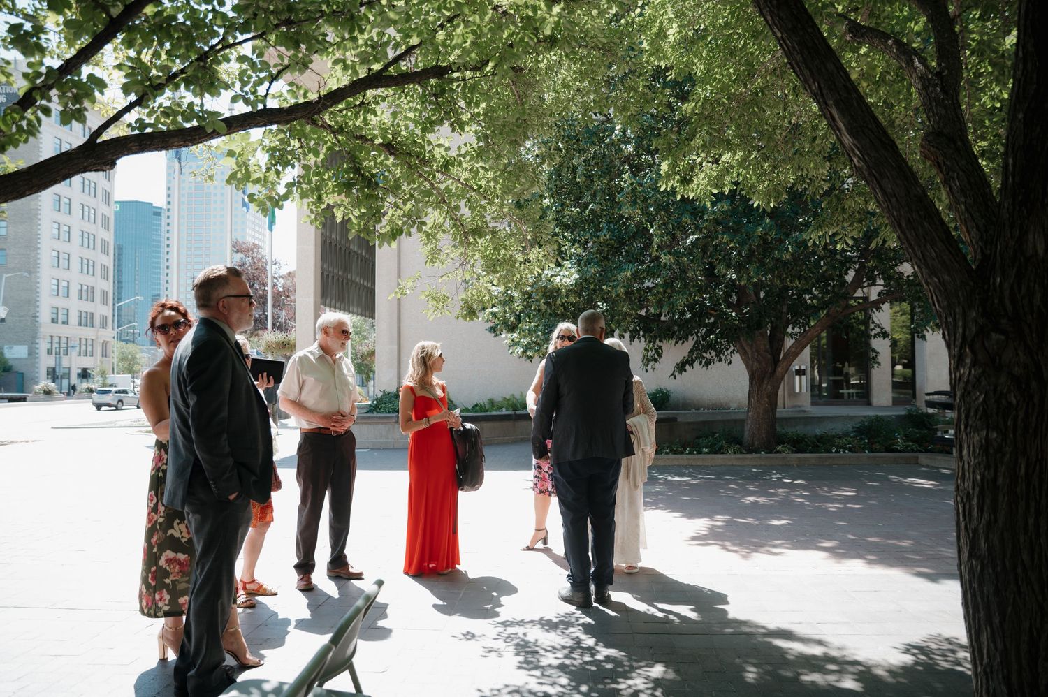 Wedding guests mingling outdoors near urban trees on a sunny day.
