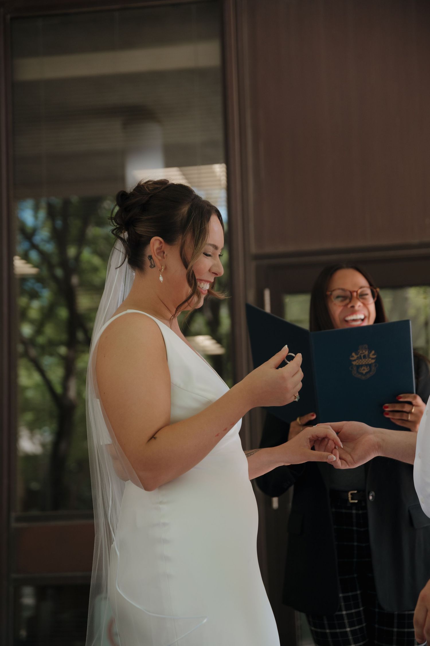Bride in white gown and veil smiling while reading vows during wedding ceremony.