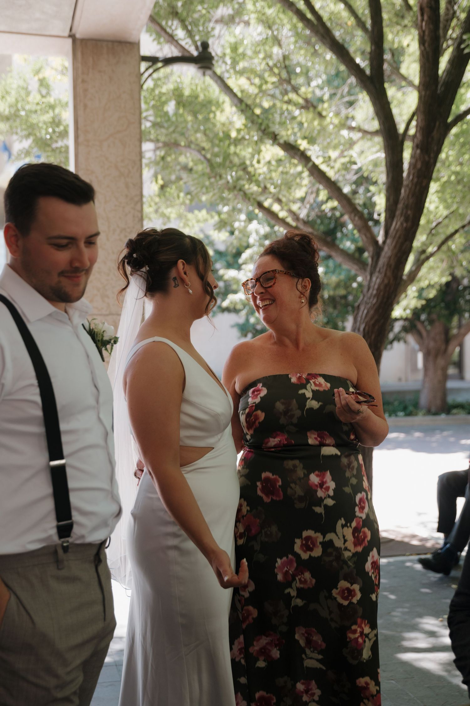 Bride and groom share a joyful moment with smiling wedding guest in floral dress.