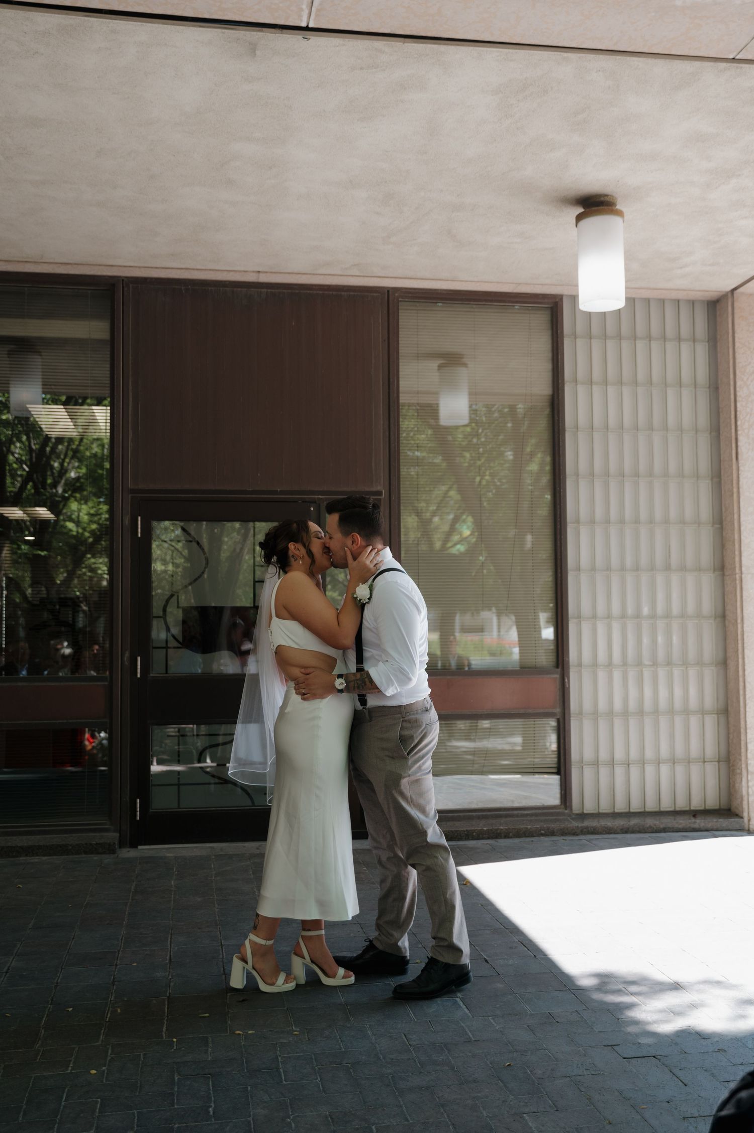 Newlywed couple shares first kiss outside modern building after wedding ceremony.