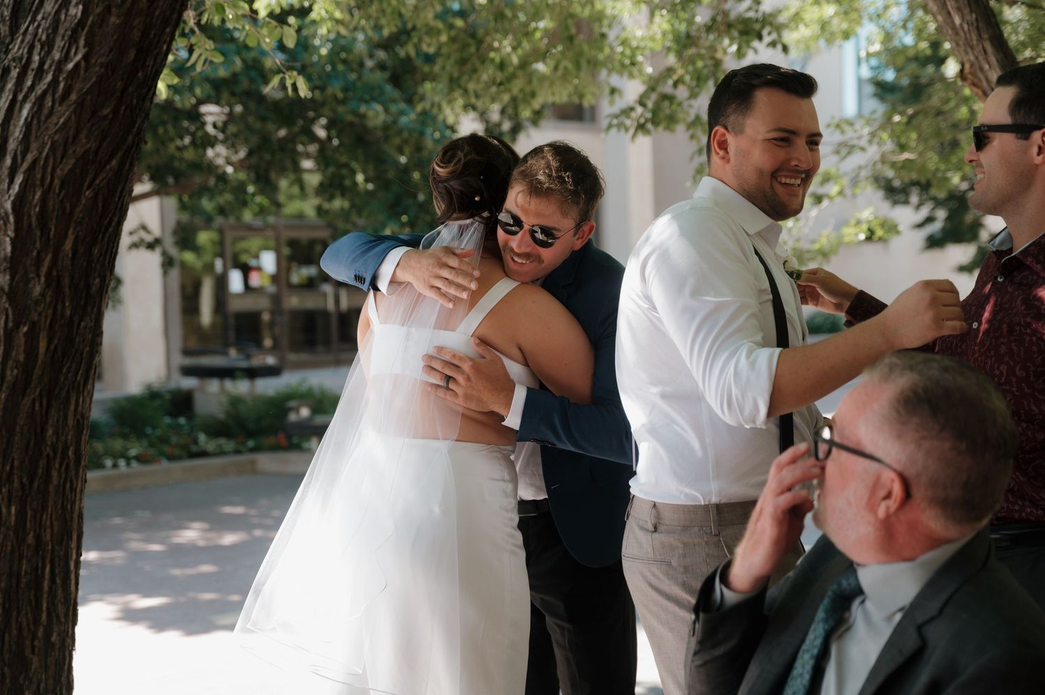 Bride in white wedding dress receives warm hugs from smiling guests at outdoor wedding reception.