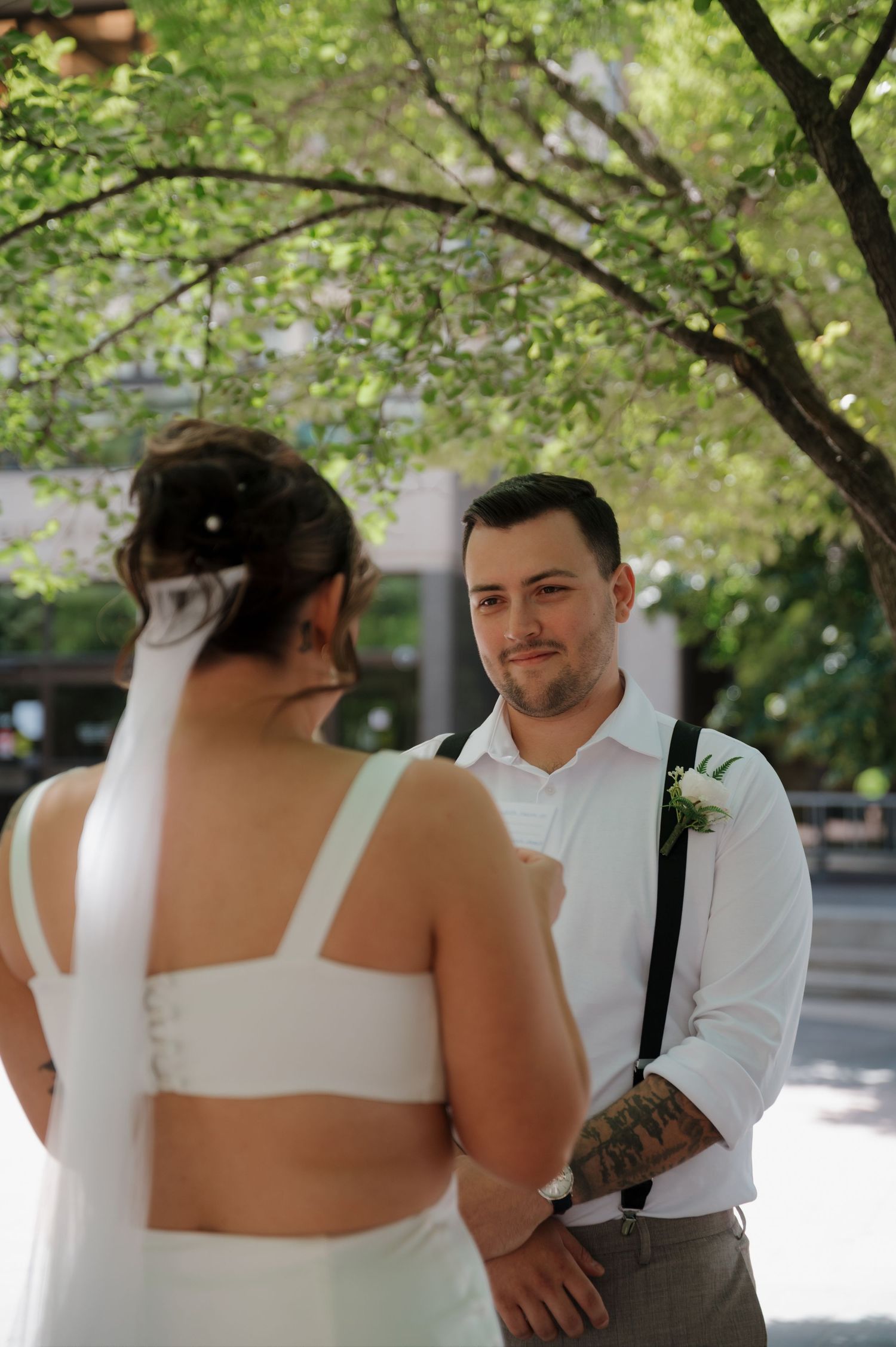Smiling groom in white shirt and suspenders gazing at bride during outdoor wedding ceremony.