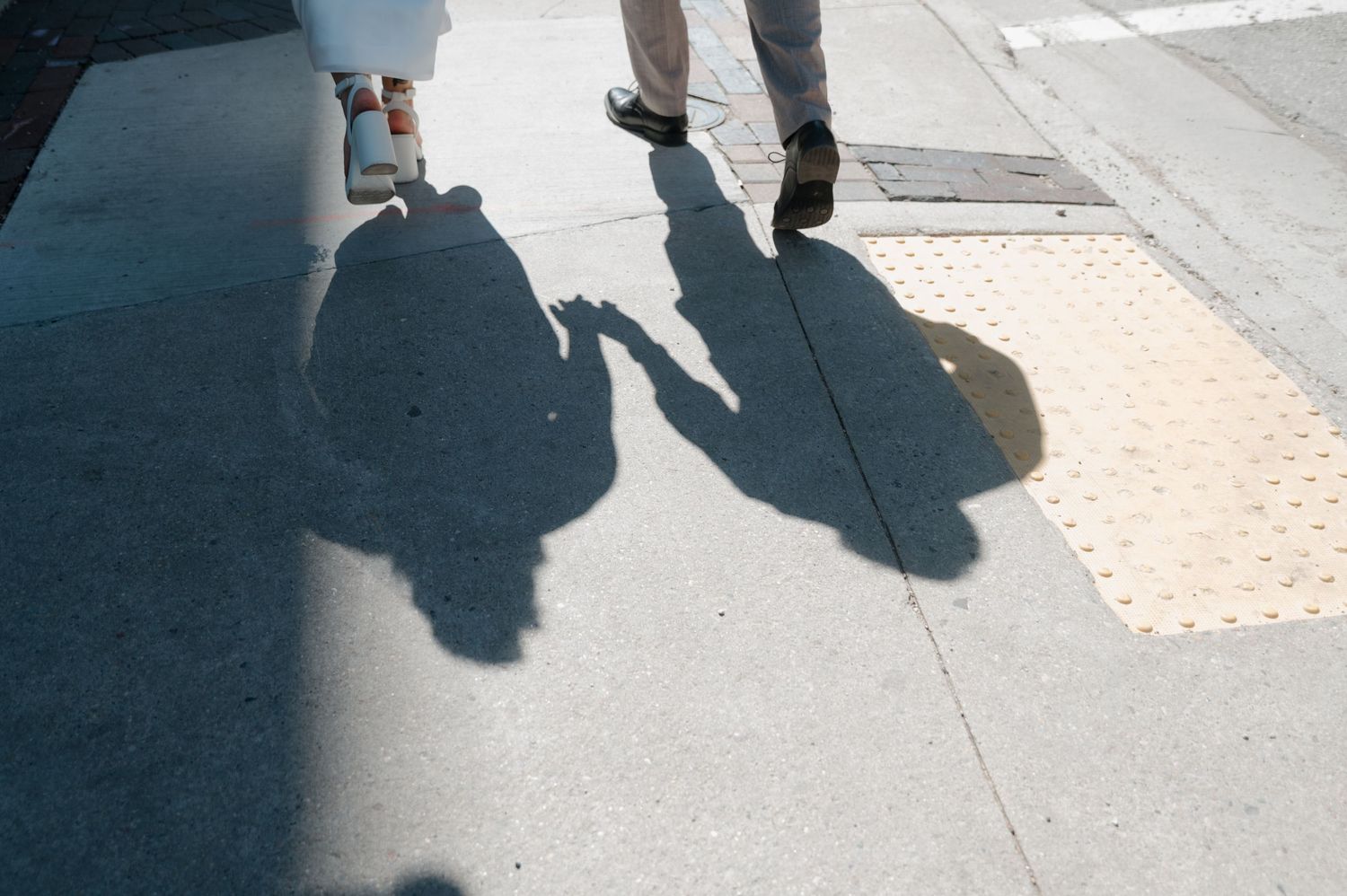 Shadows of newlywed couple holding hands cast on sunny sidewalk pavement.