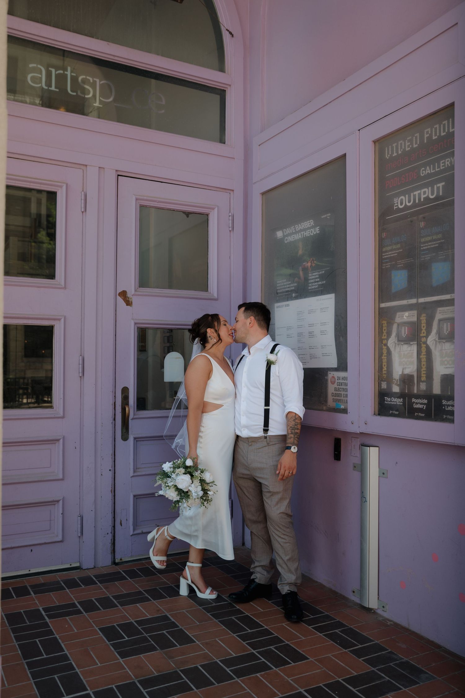 Bride and groom kissing outside colorful Artspace gallery entrance with wedding bouquet.