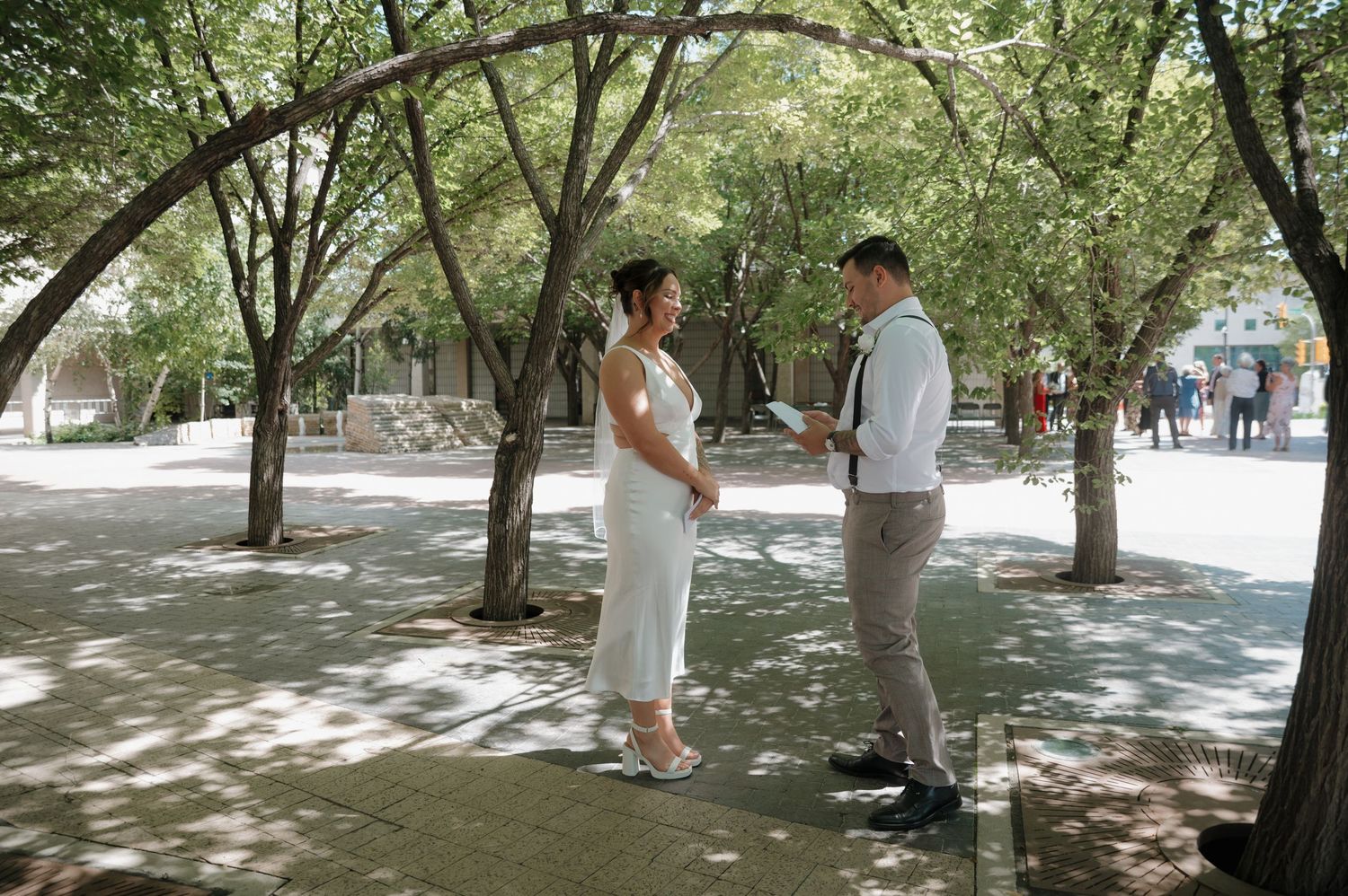 Bride and groom sharing a moment under arched trees in a sunny outdoor setting.