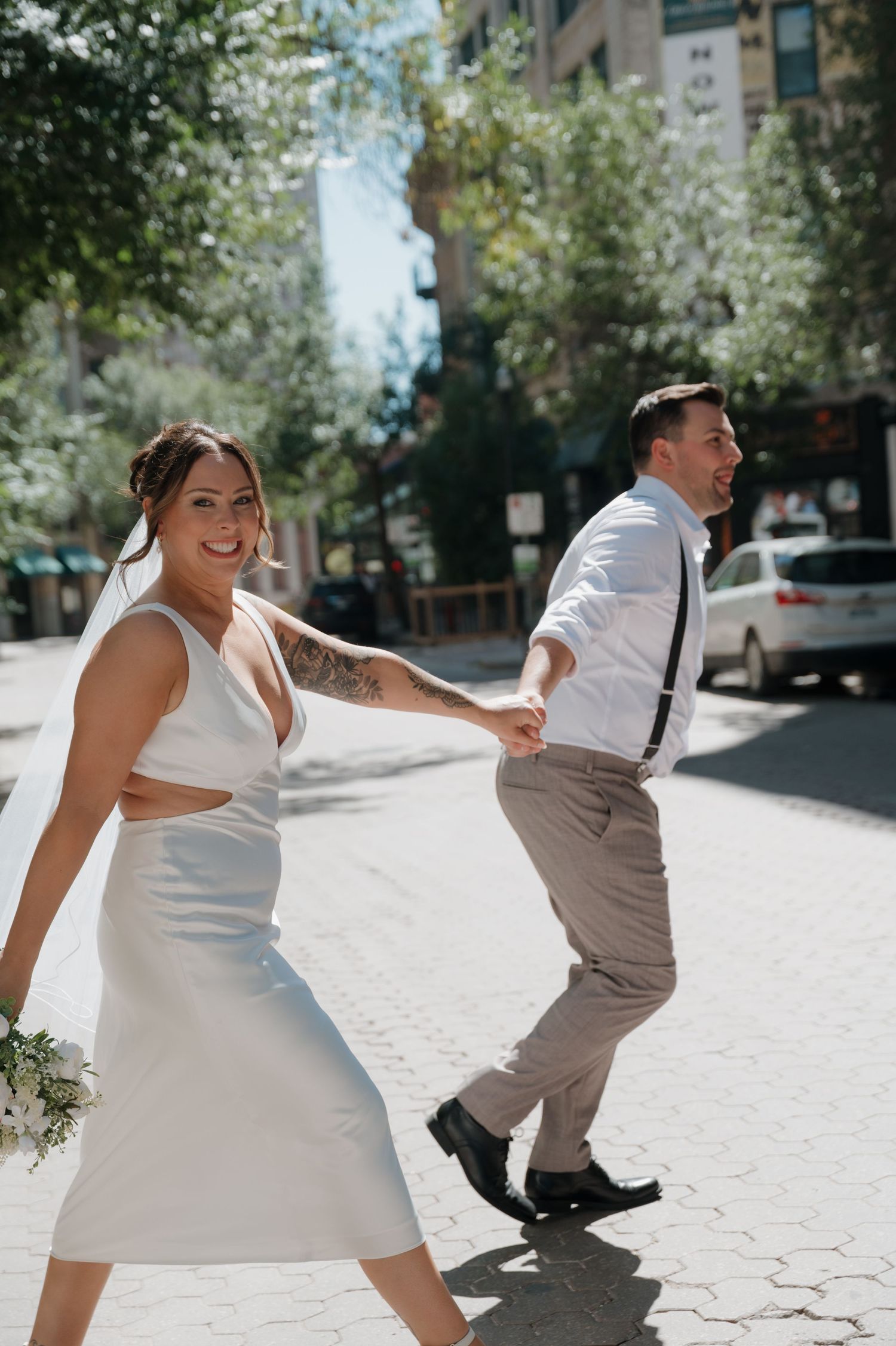 Smiling bride in white midi dress holding bouquet walks hand-in-hand with groom on a sunny city street.
