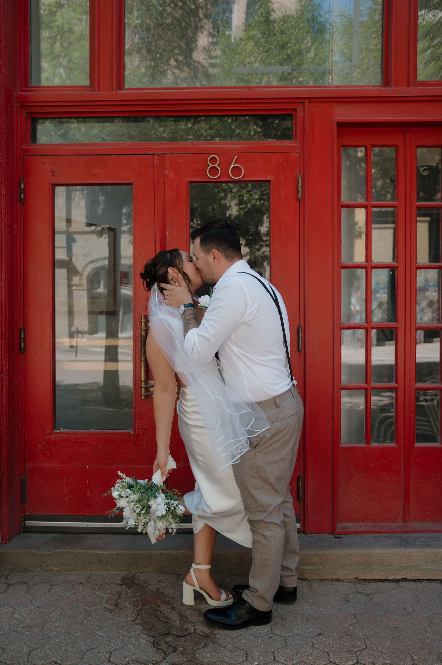 Bride and groom sharing passionate kiss in front of bold red doors numbered 86.