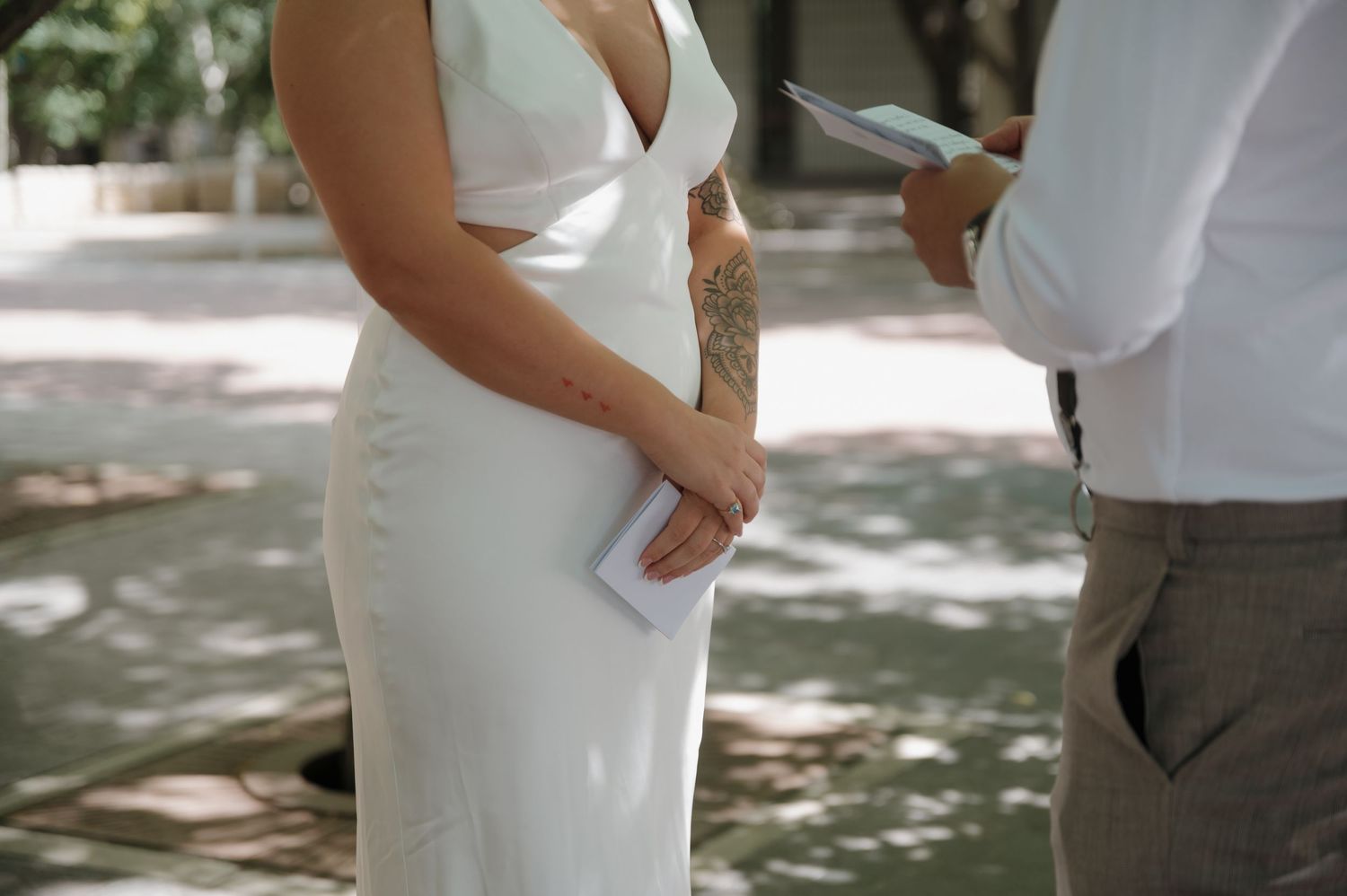 Bride in a white sleeveless wedding gown with cutout detail holding vows during an outdoor ceremony.