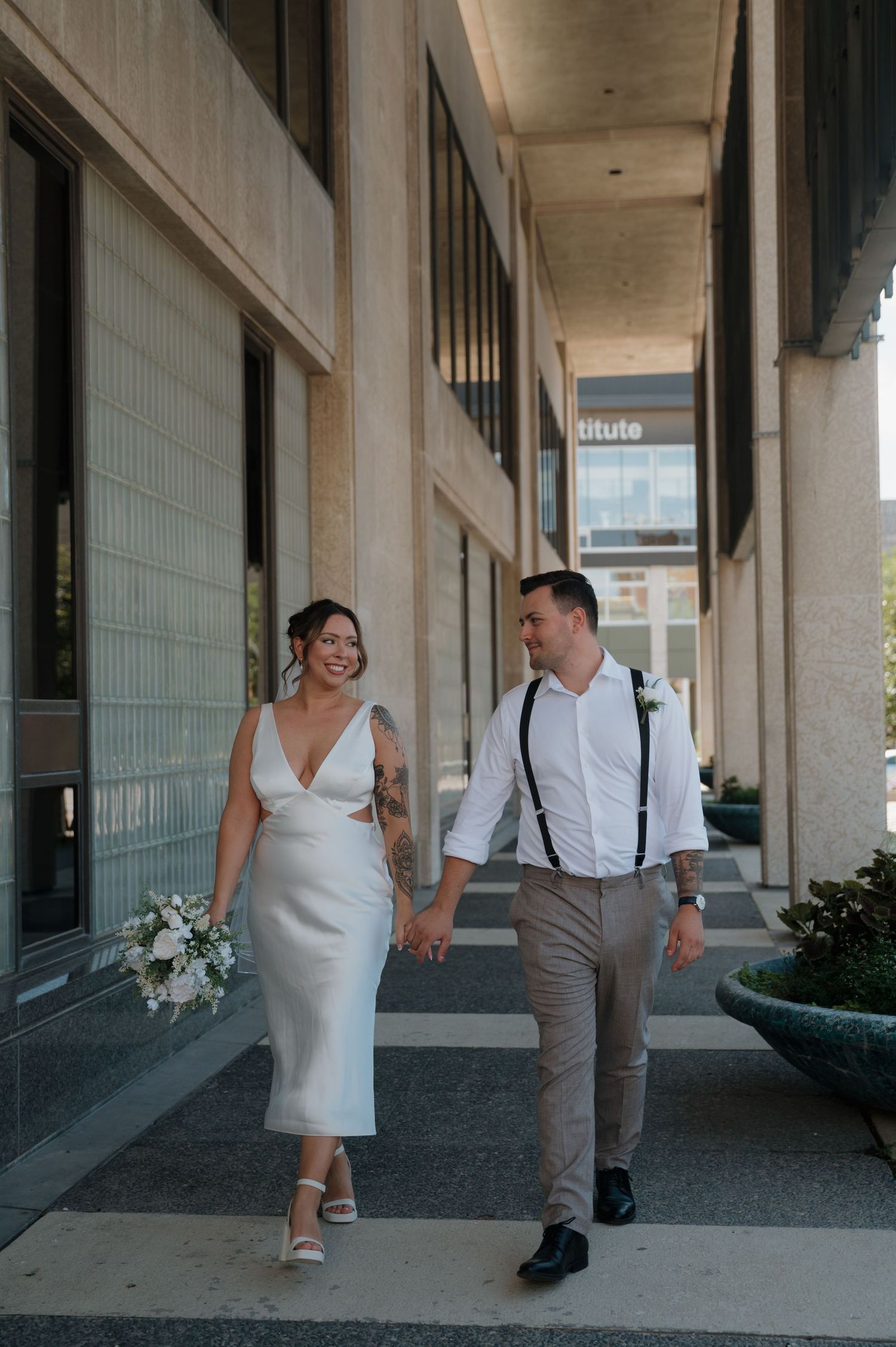 Bride and groom walking hand in hand, bride in white midi dress holding bouquet, groom in suspenders.