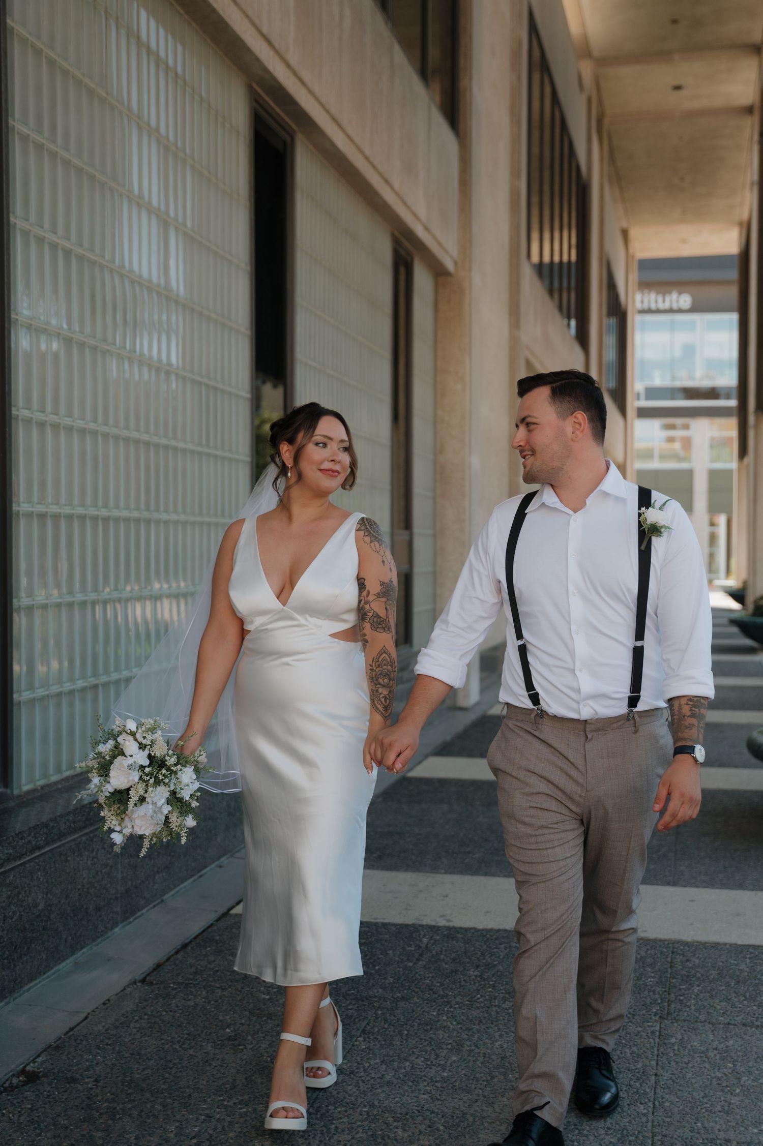 Bride and groom walking hand in hand, bride wearing white midi dress with bouquet.