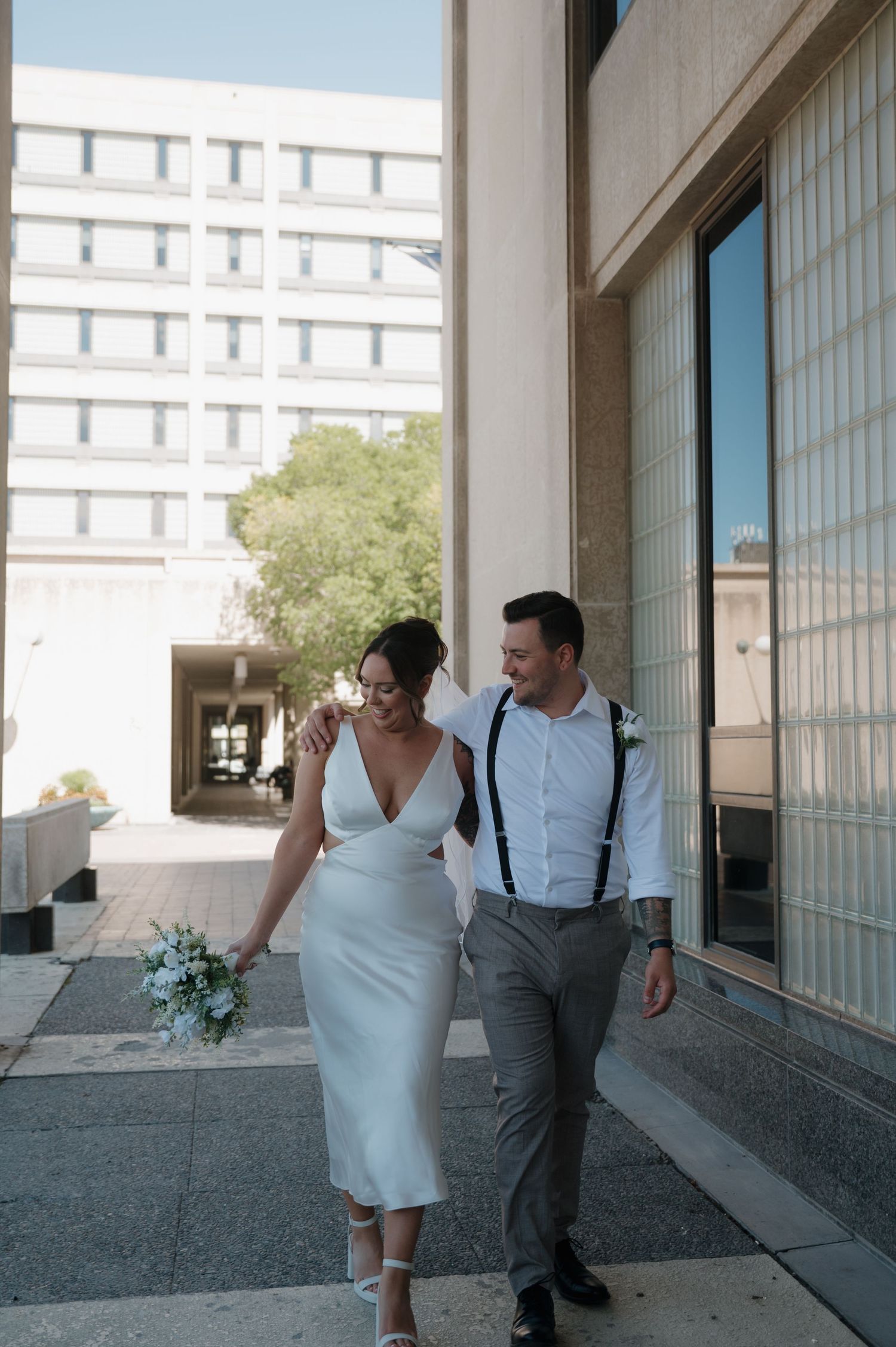 Bride and groom walking together outside modern building after wedding ceremony.