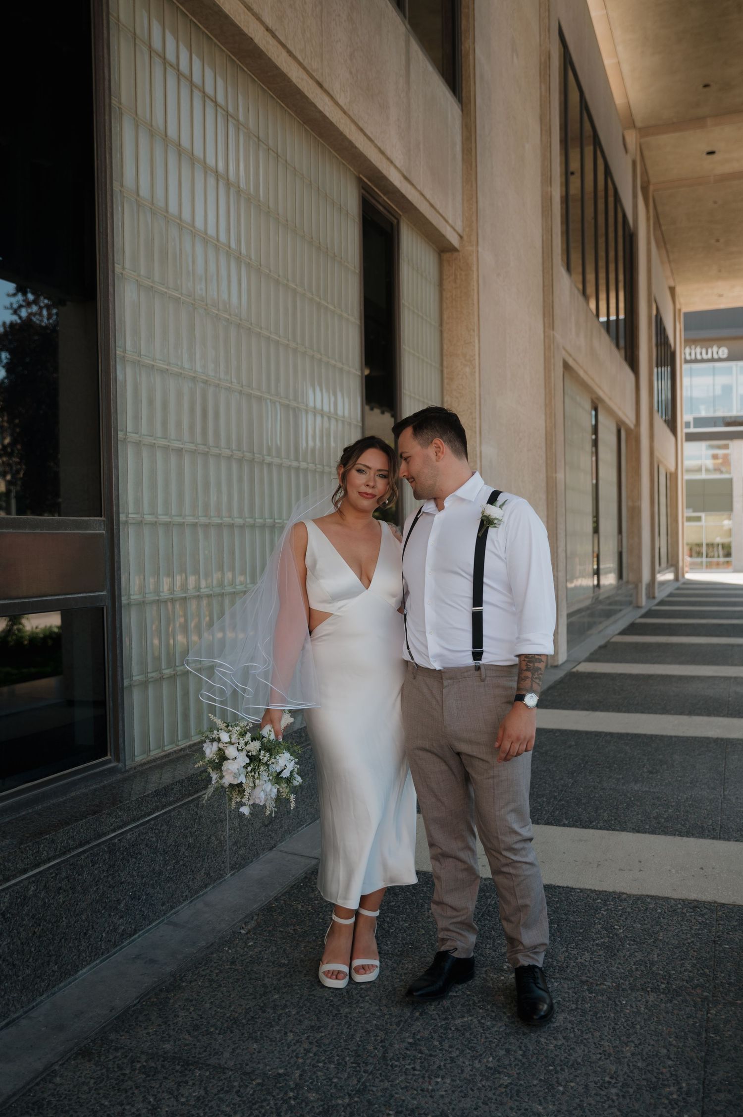 Bride in white midi dress holding bouquet leans on groom in white shirt with suspenders outside modern building.