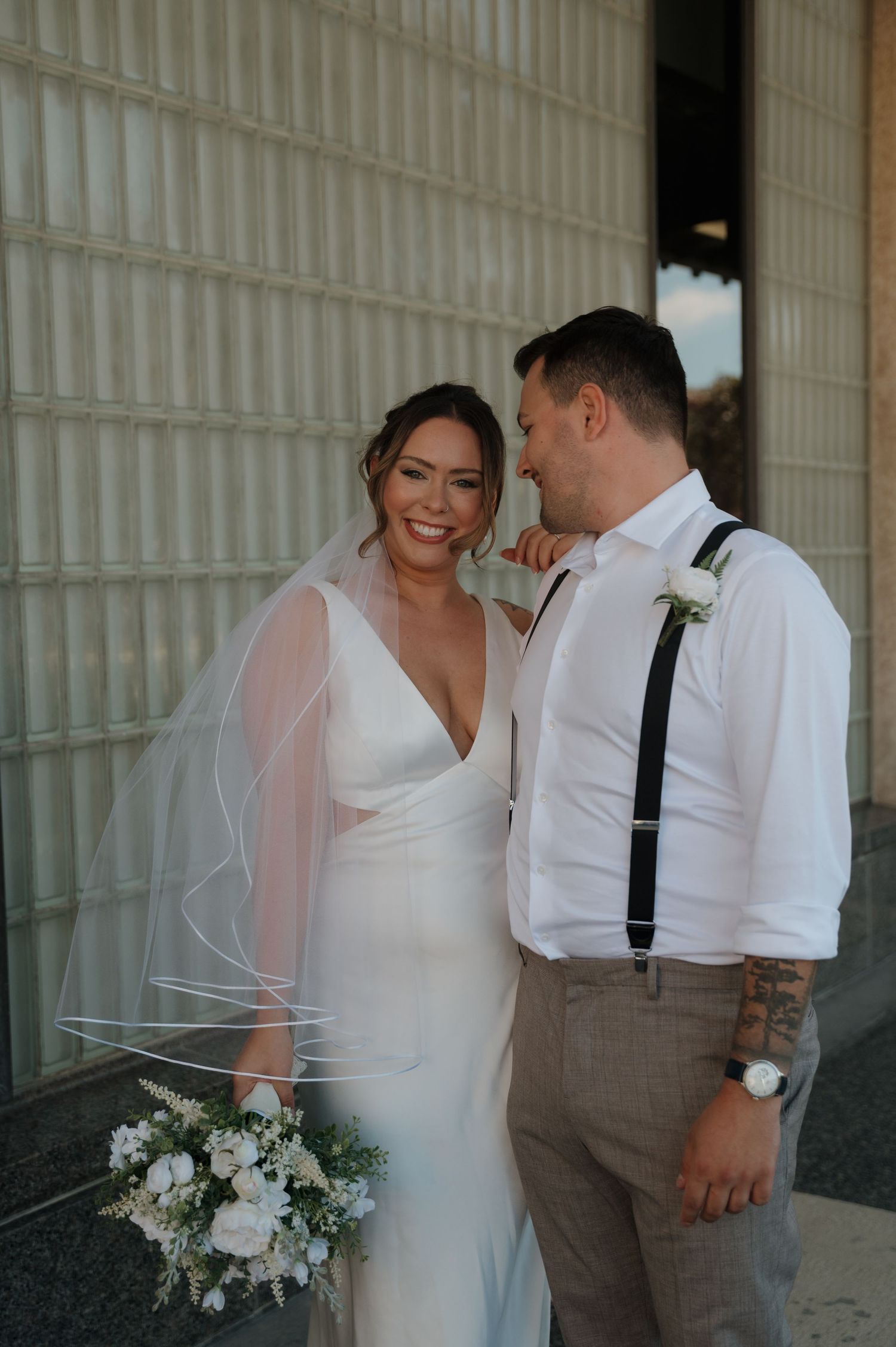 Happy bride in white gown with veil holding bouquet stands with groom in suspenders outdoors.