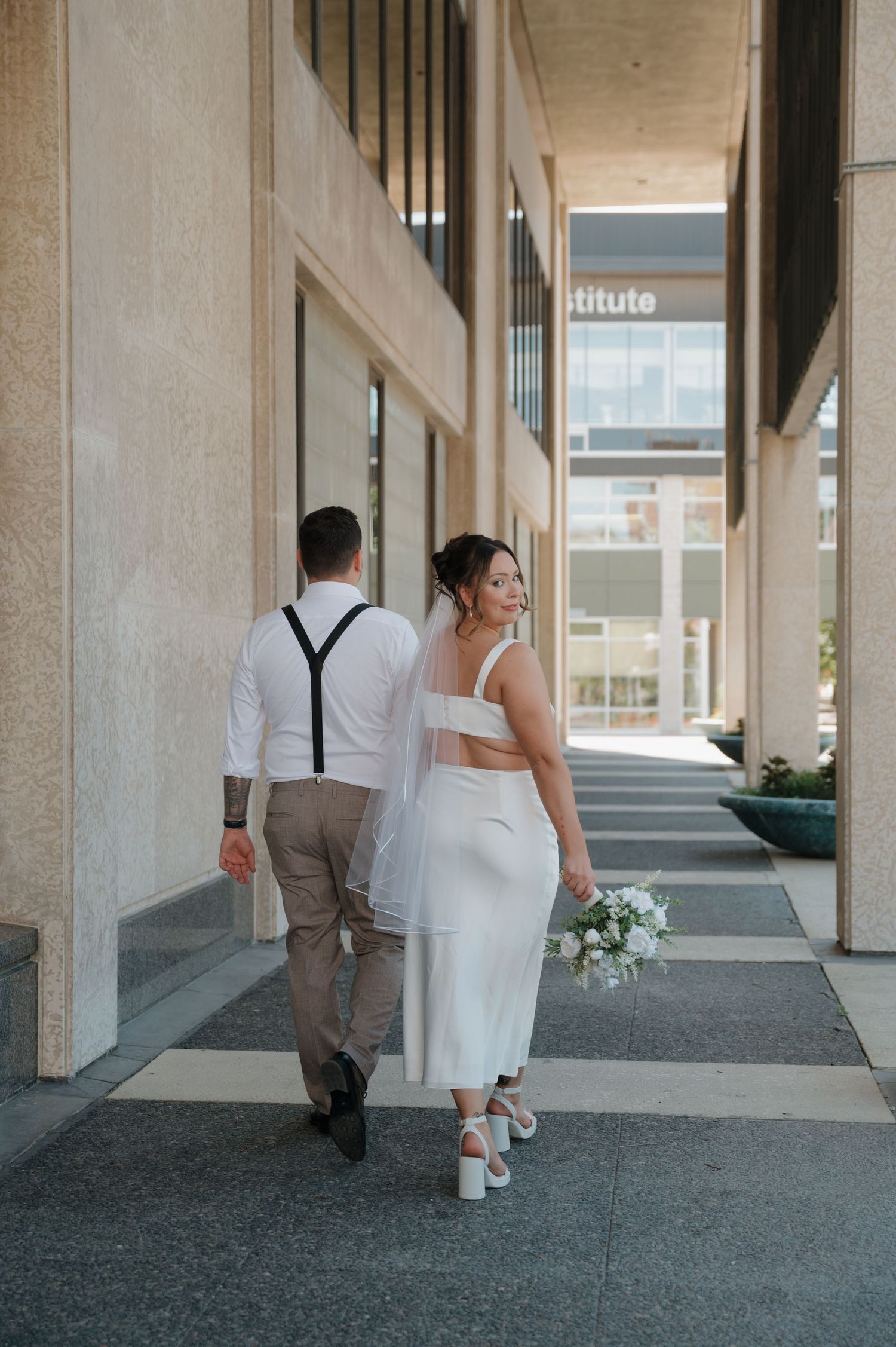 Bride in white crop top skirt holding bouquet walks with groom in suspenders along modern building.