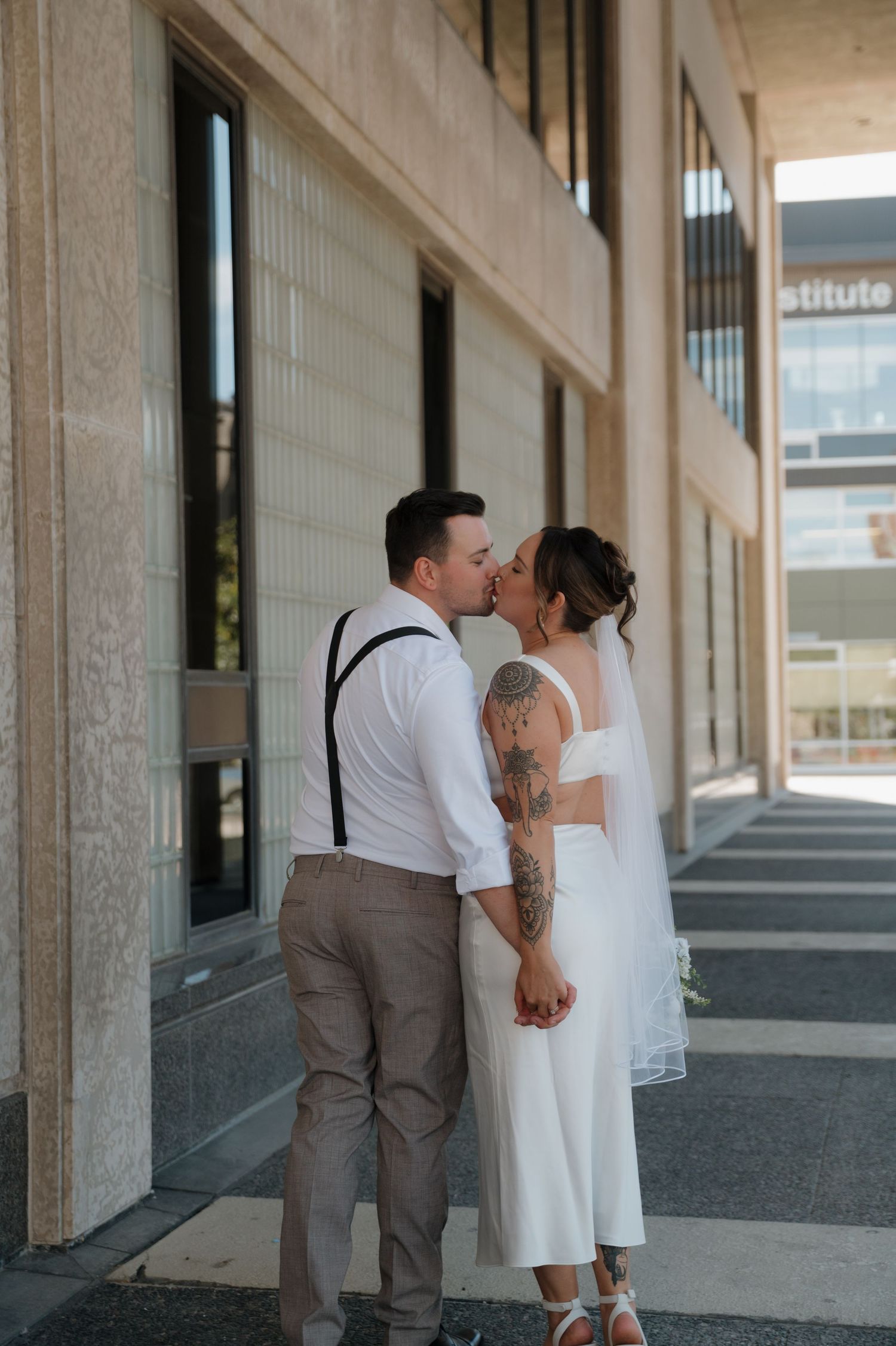 Bride and groom sharing a kiss outside modern building, bride in white dress with veil and tattoos.