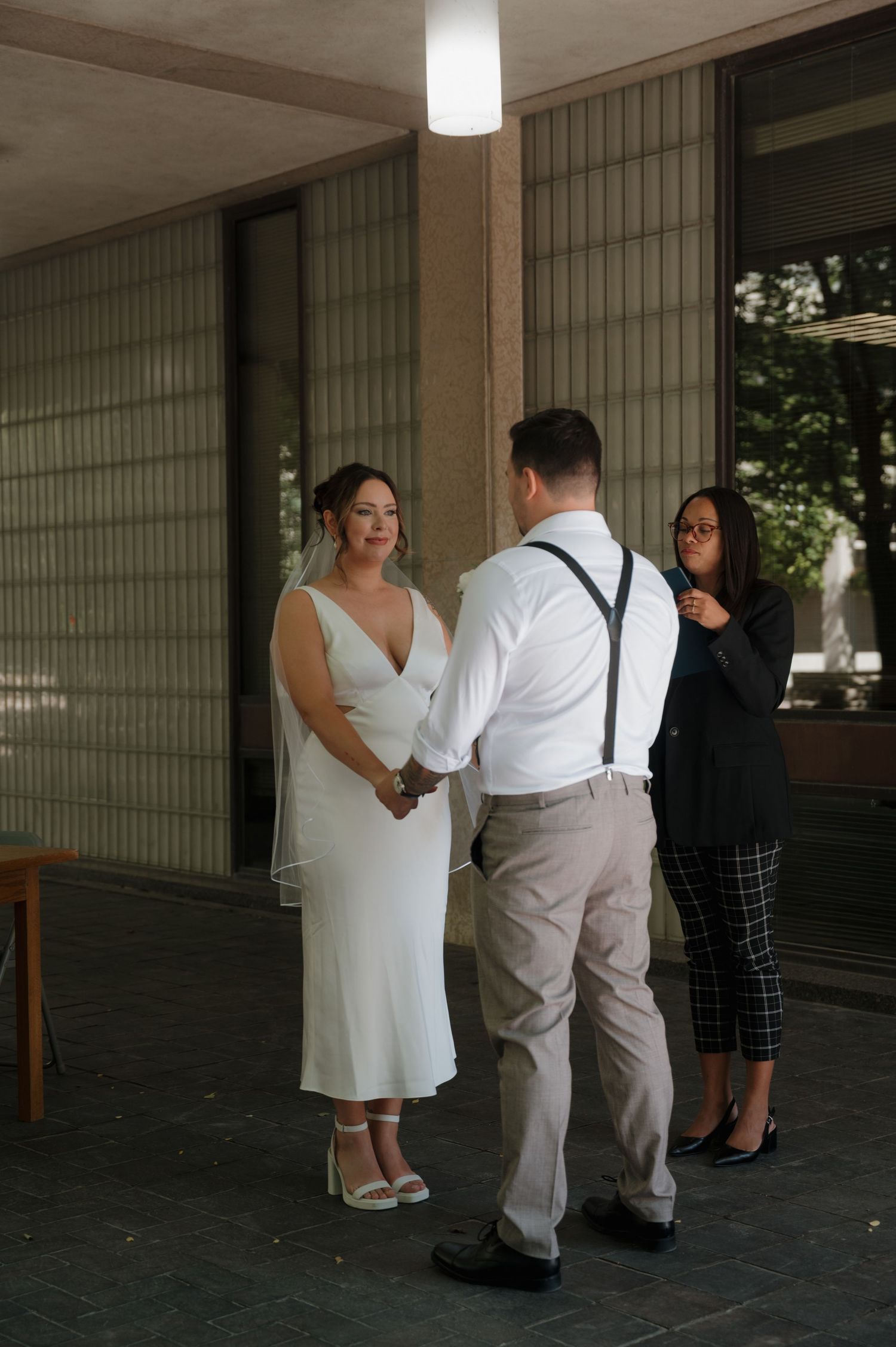 Bride in white dress and groom in suspenders hold hands during wedding ceremony.