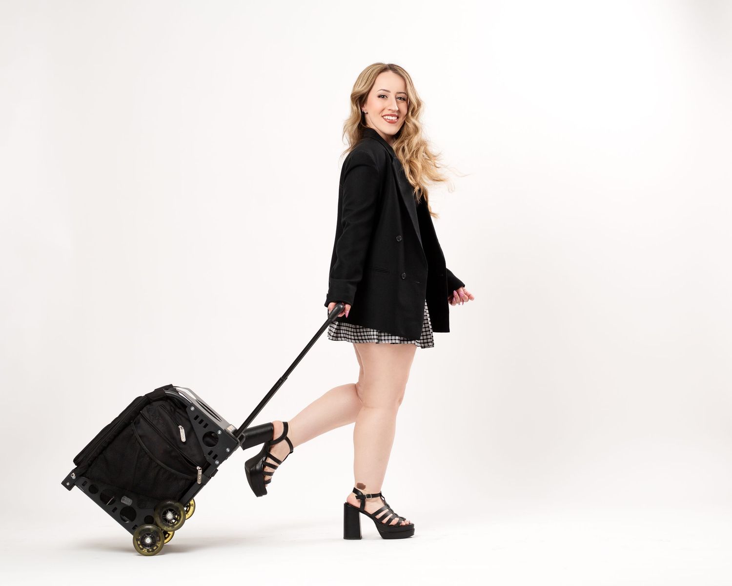 Stylish traveler in black blazer and dress pulling wheeled luggage against white background.