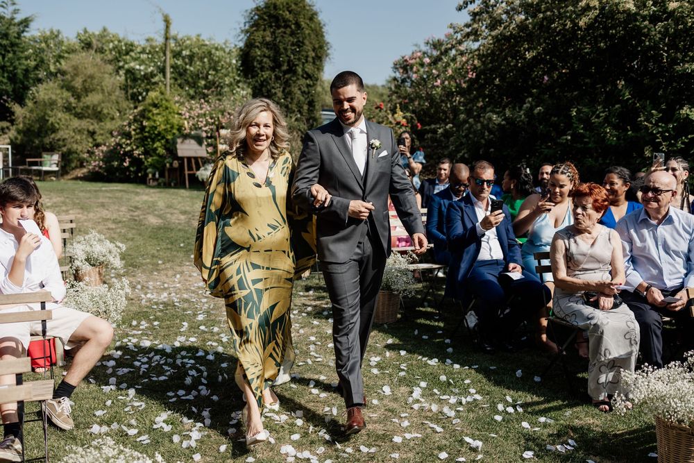 Wedding guests gather outdoors on a sunny day in a natural setting with scattered white petals on the ground.