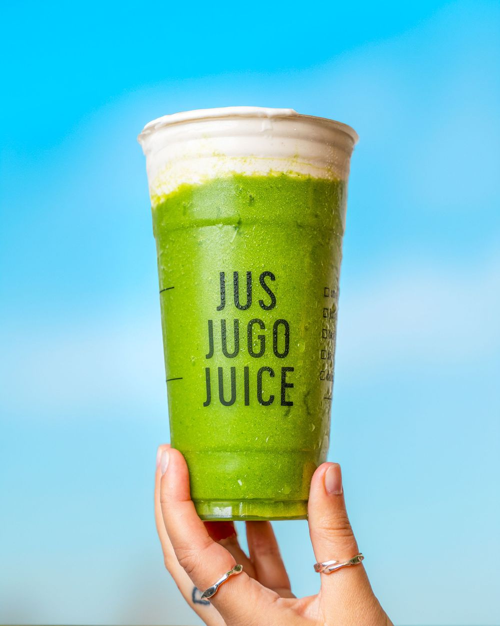 Hand holding a green smoothie in a clear plastic cup with 'JUS JUGO JUICE' text against a bright blue sky background.