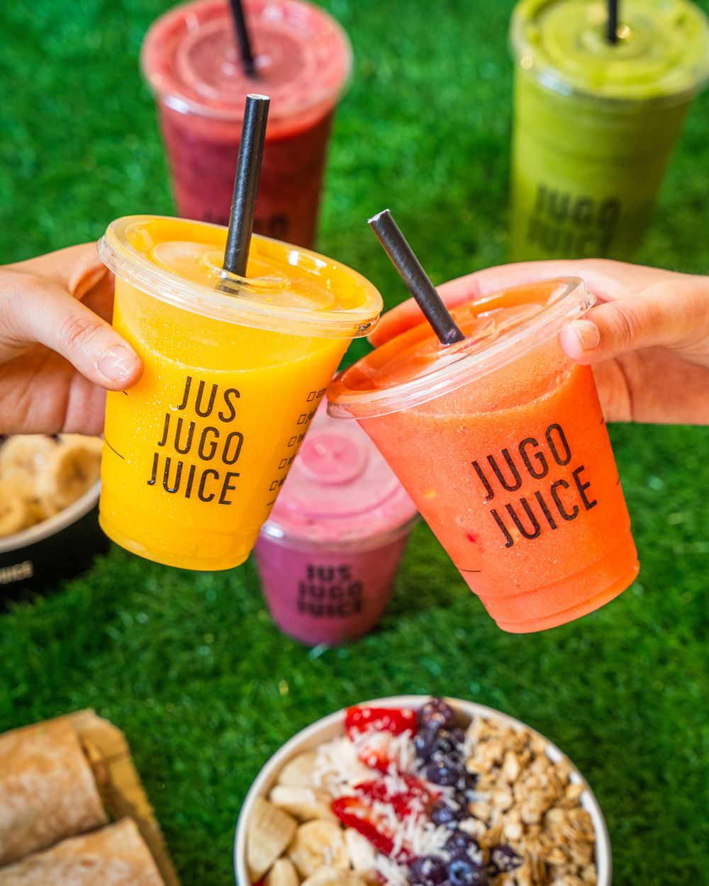 Colorful smoothies in plastic cups with straws arranged on grass next to acai bowls topped with fresh fruit.