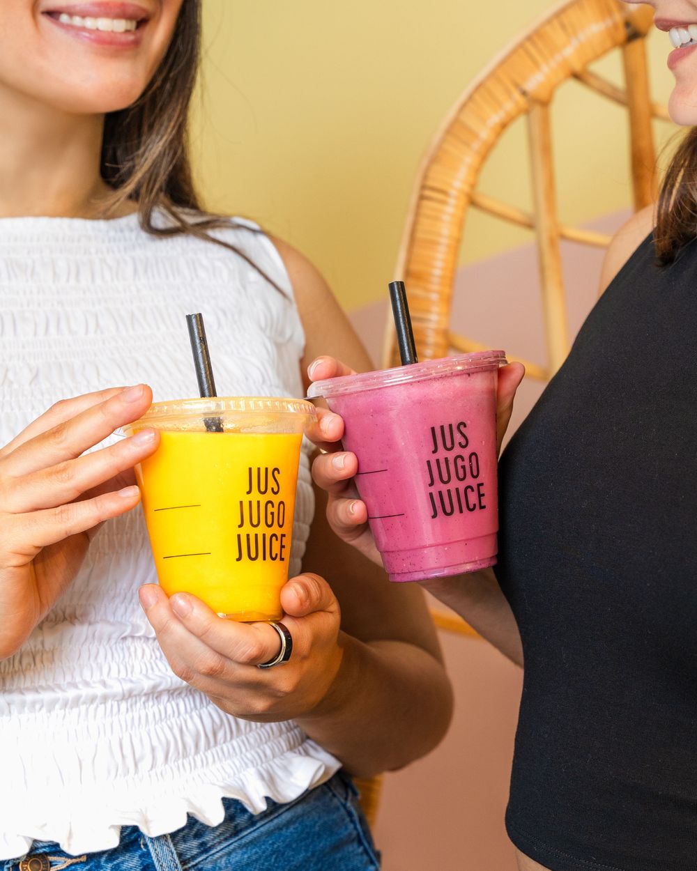 Two colorful smoothies in plastic cups from Jus Jugo Juice being held by people wearing casual clothing.