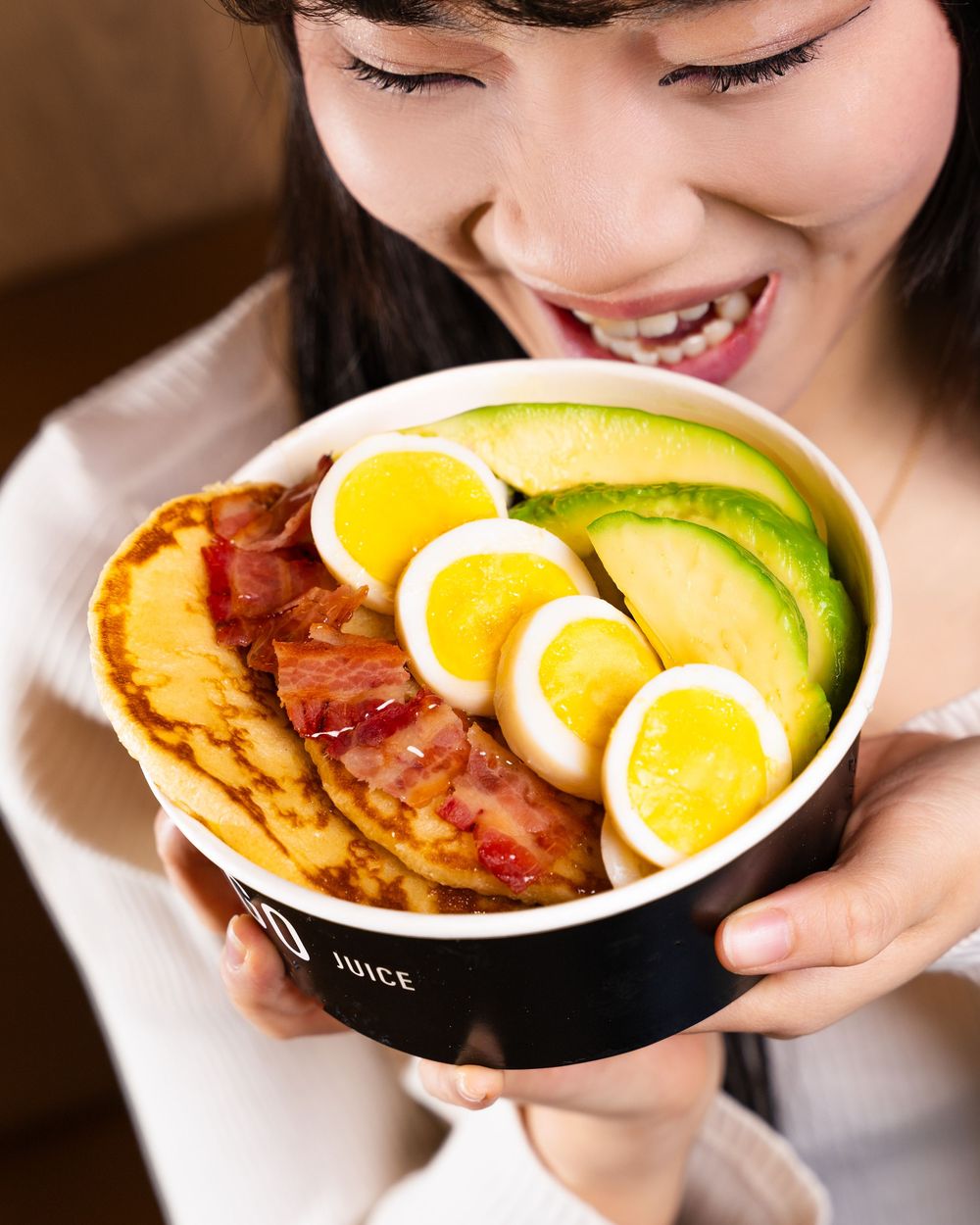 A breakfast bowl filled with sliced avocado, boiled eggs, and bacon served in a black container.
